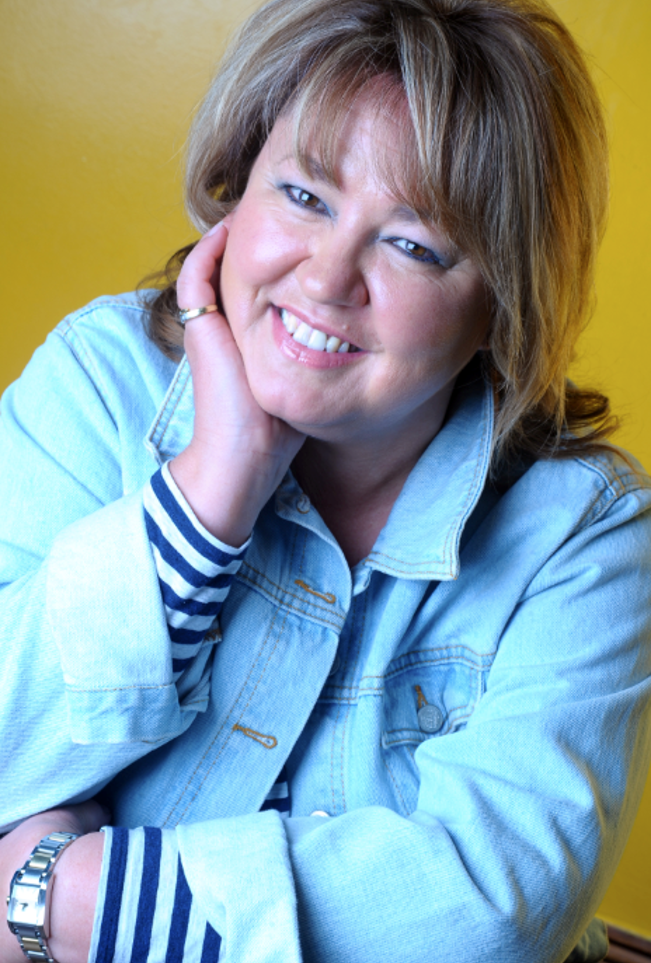 A woman smiling, wearing a light blue denim jacket over a striped shirt, with her hand resting on the side of her face, set against a yellow background.