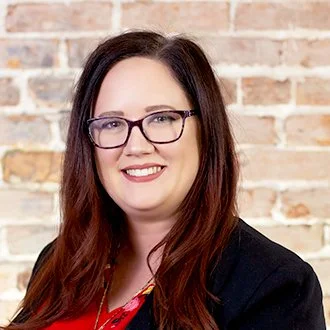 A woman with long brown hair and glasses smiling in front of a brick wall.