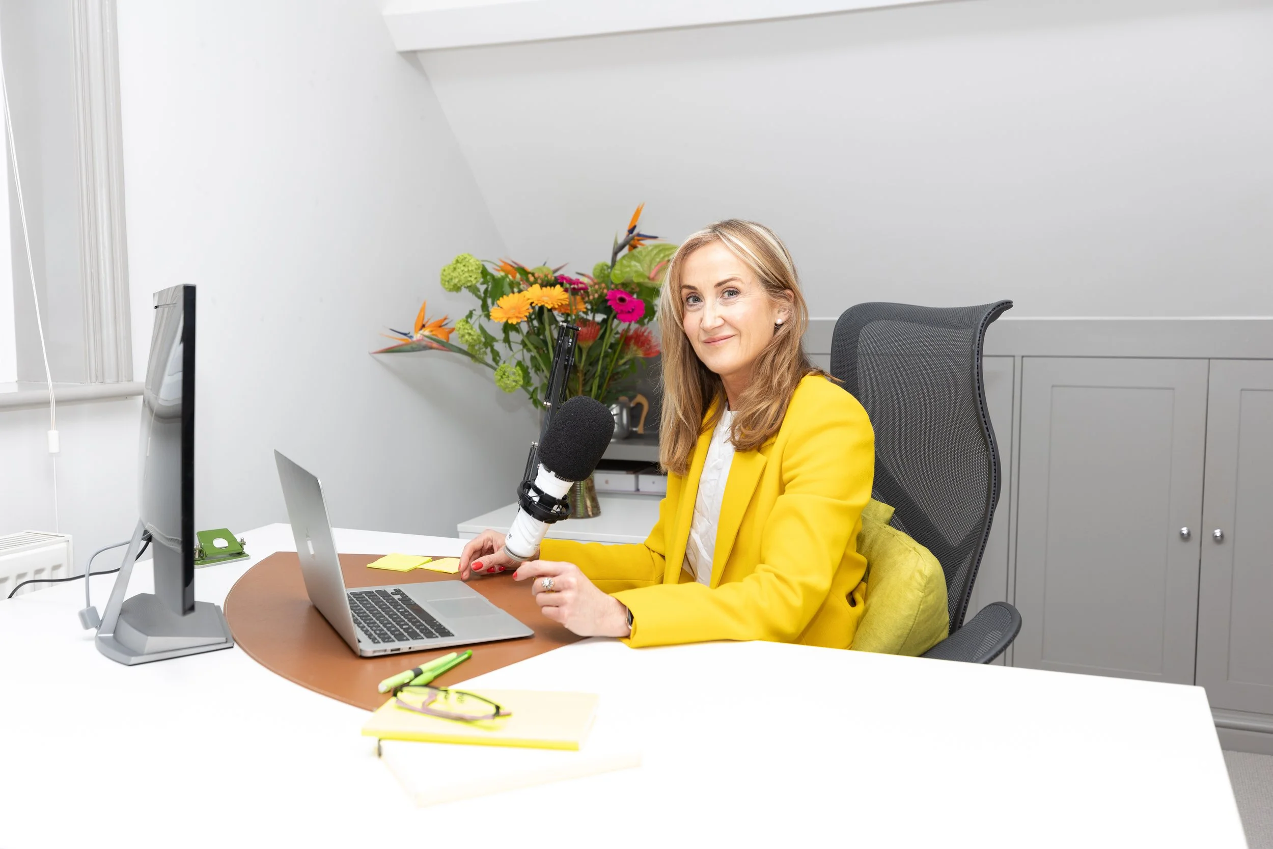 A woman in a yellow blazer sitting at a desk with a laptop, microphone, flowers, and office supplies.