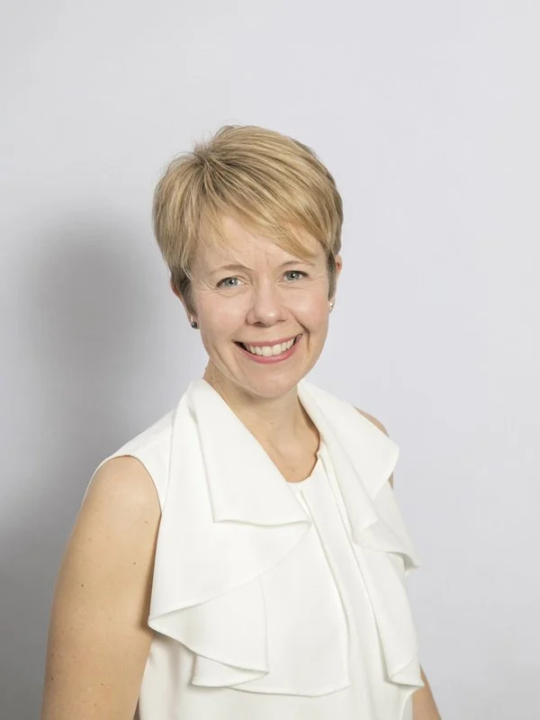 A woman with short blonde hair smiling, wearing a white sleeveless top, against a plain light gray background.