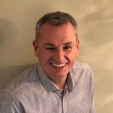 A smiling man with short gray hair wearing a light-colored patterned shirt.