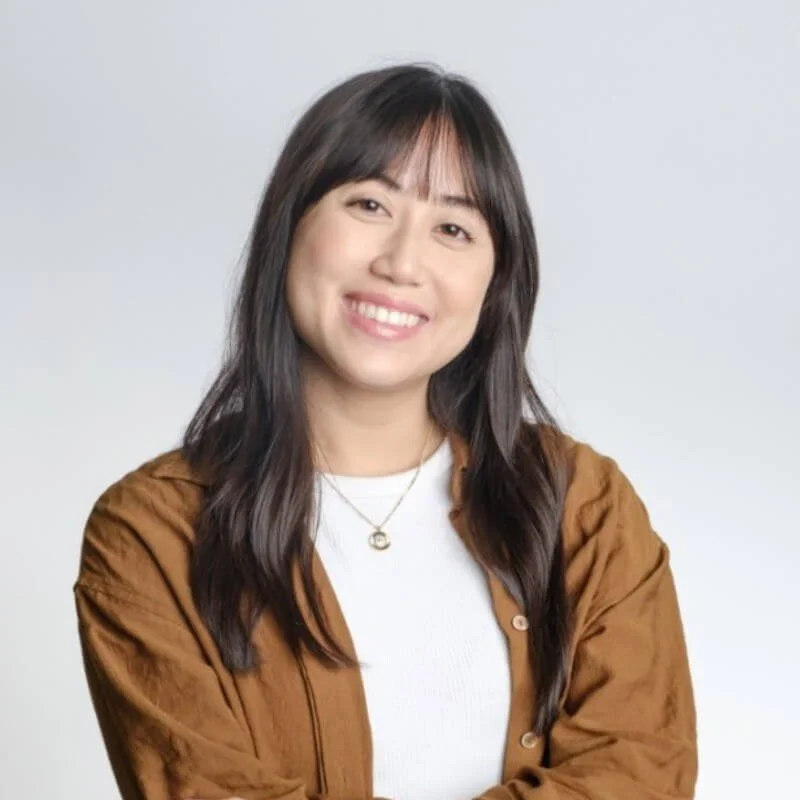 A young Asian woman with long dark hair, smiling, wearing a white shirt and a brown jacket, against a plain light gray background.