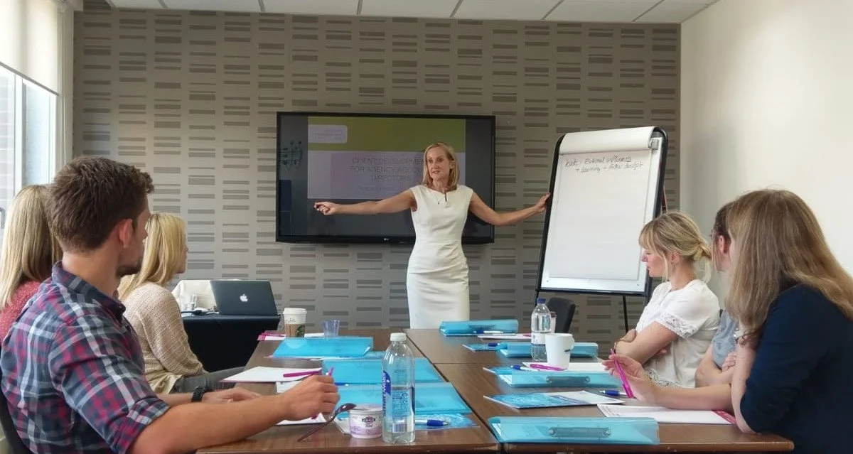 Businesswoman giving a presentation to a group of six people in a conference room with a large screen, flipboard, laptops, notebooks, and water bottles on the table.