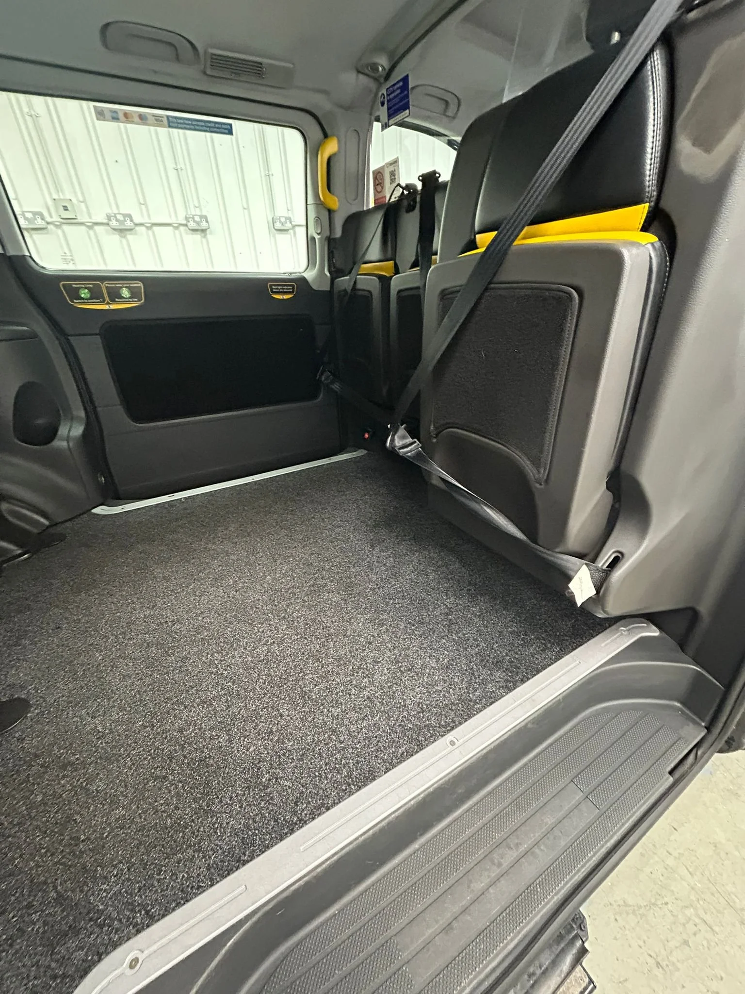 Interior view of the rear passenger area of a vehicle with the seats folded forward, showing a black carpeted floor and black seats with yellow accents.