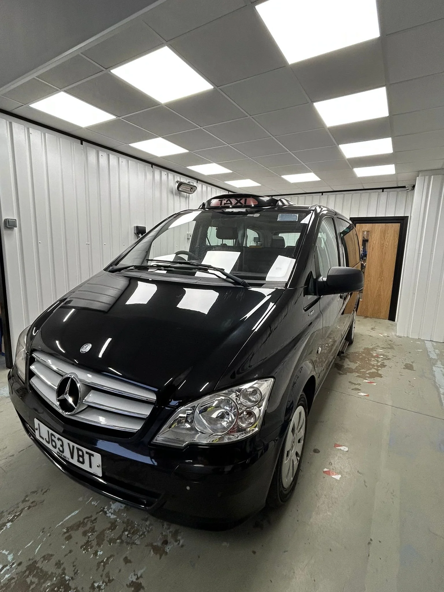 Black Mercedes-Benz taxi van parked indoors, with a Taxi sign on the roof, in a brightly lit room with white paneled walls and a wooden door in the background.