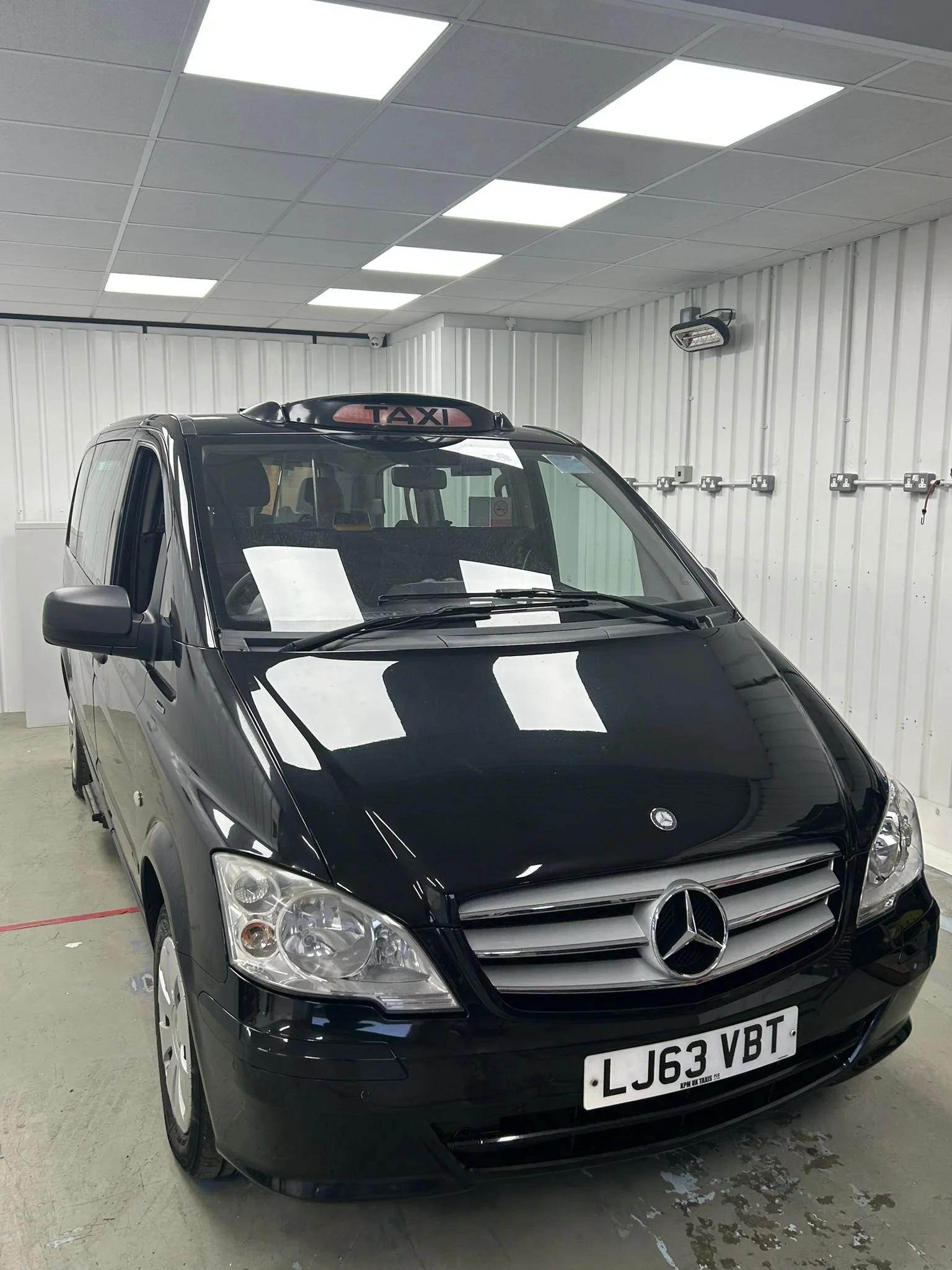 Black Mercedes-Benz taxi van with a taxi sign on the roof inside a garage, showing front view.