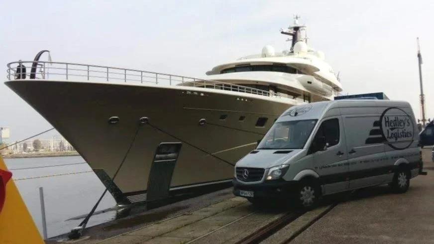 Large yacht docked at the pier next to a logistics van on a road near water.