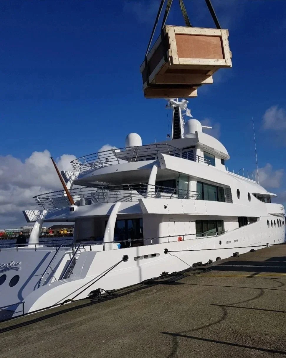 Large white yacht docked at a pier, with a crane lifting a wooden crate above it against a blue sky with clouds.