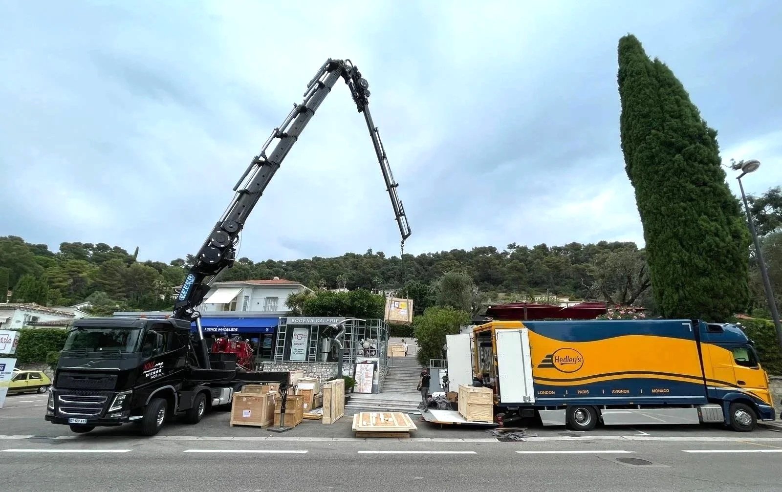 A crane truck unloading wooden crates in front of a building, with another delivery truck parked nearby, and a tall green tree in the background.
