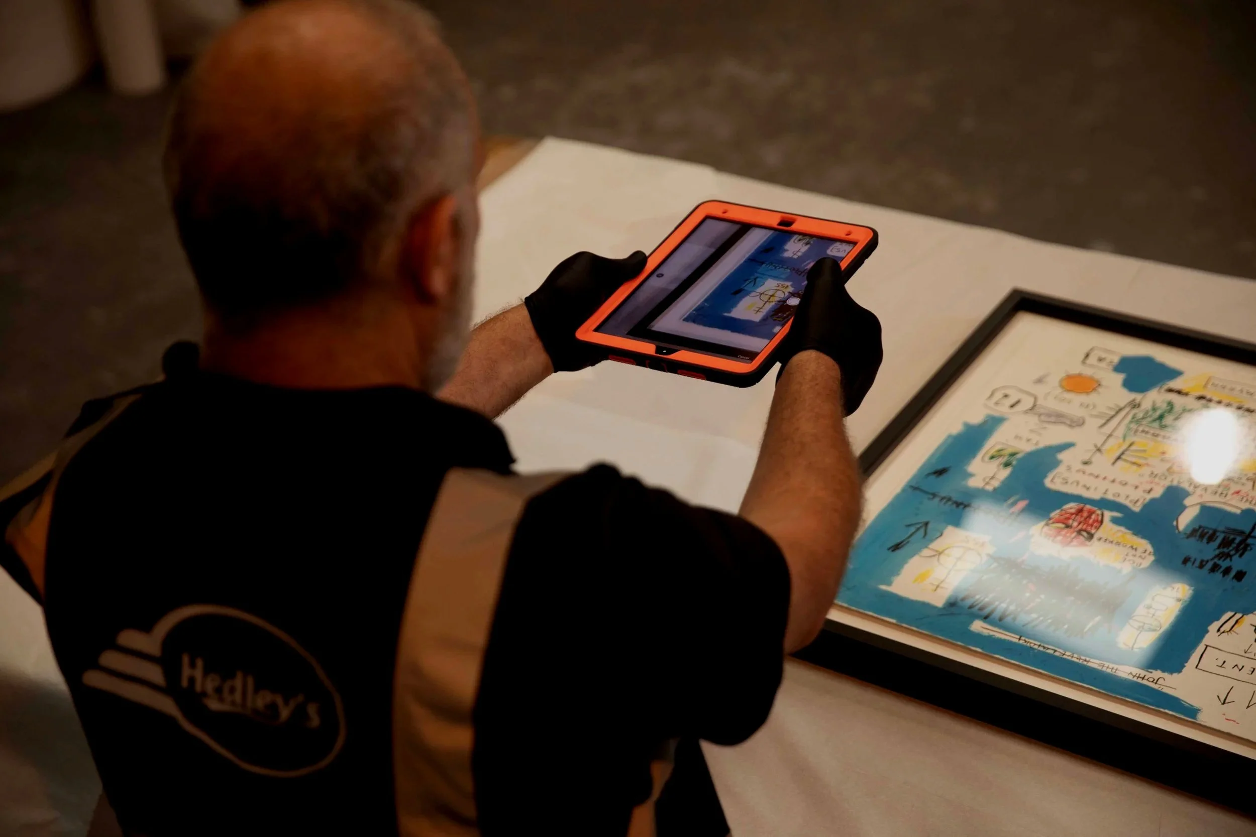 An older man with a beard and glasses, wearing gloves and a black and tan shirt with a Hedley's logo, is using a tablet to look at a framed artwork on a table. The artwork appears to be a colorful, hand-drawn map or illustration.