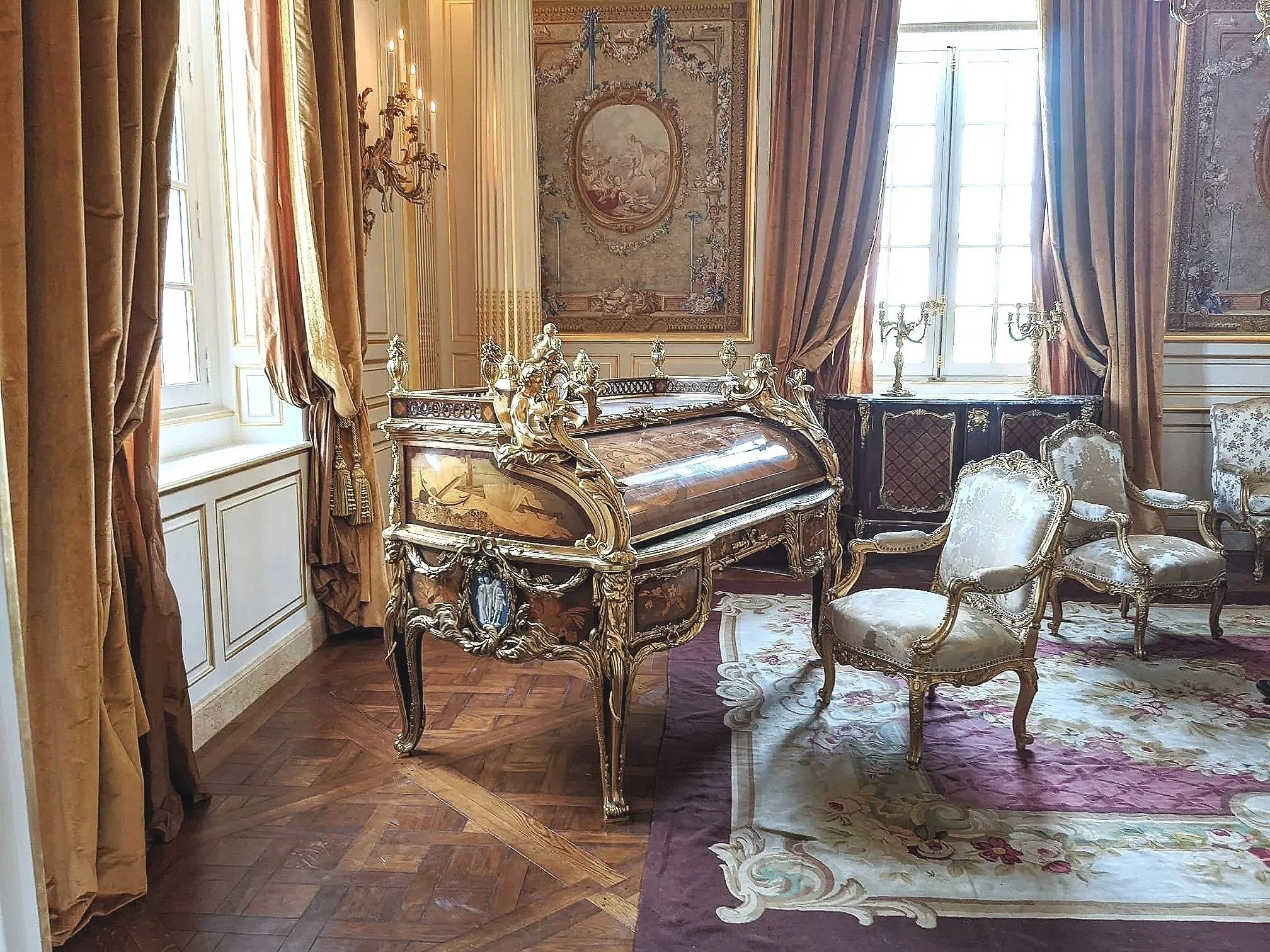 A luxurious room with ornate furniture and decor, including a decorated desk, a patterned area rug, and upholstered chairs, with large windows and gold curtains.
