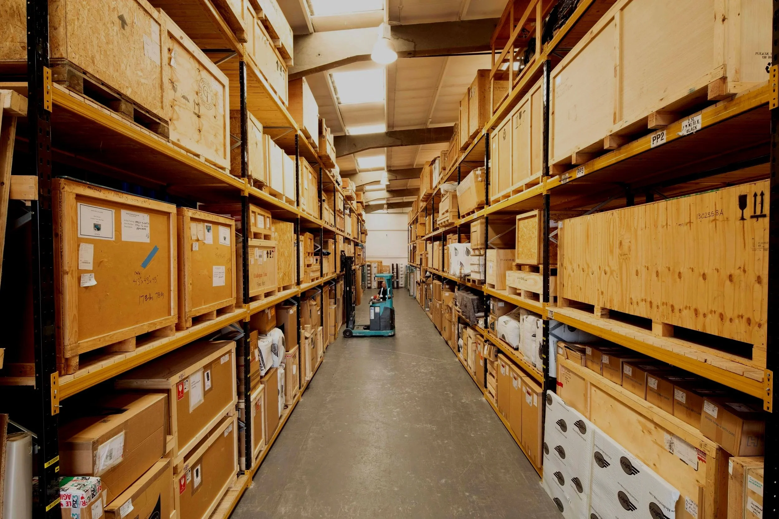 A warehouse aisle with tall metal shelves filled with large wooden crates and boxes, a green hand truck in the middle, and fluorescent ceiling lights.