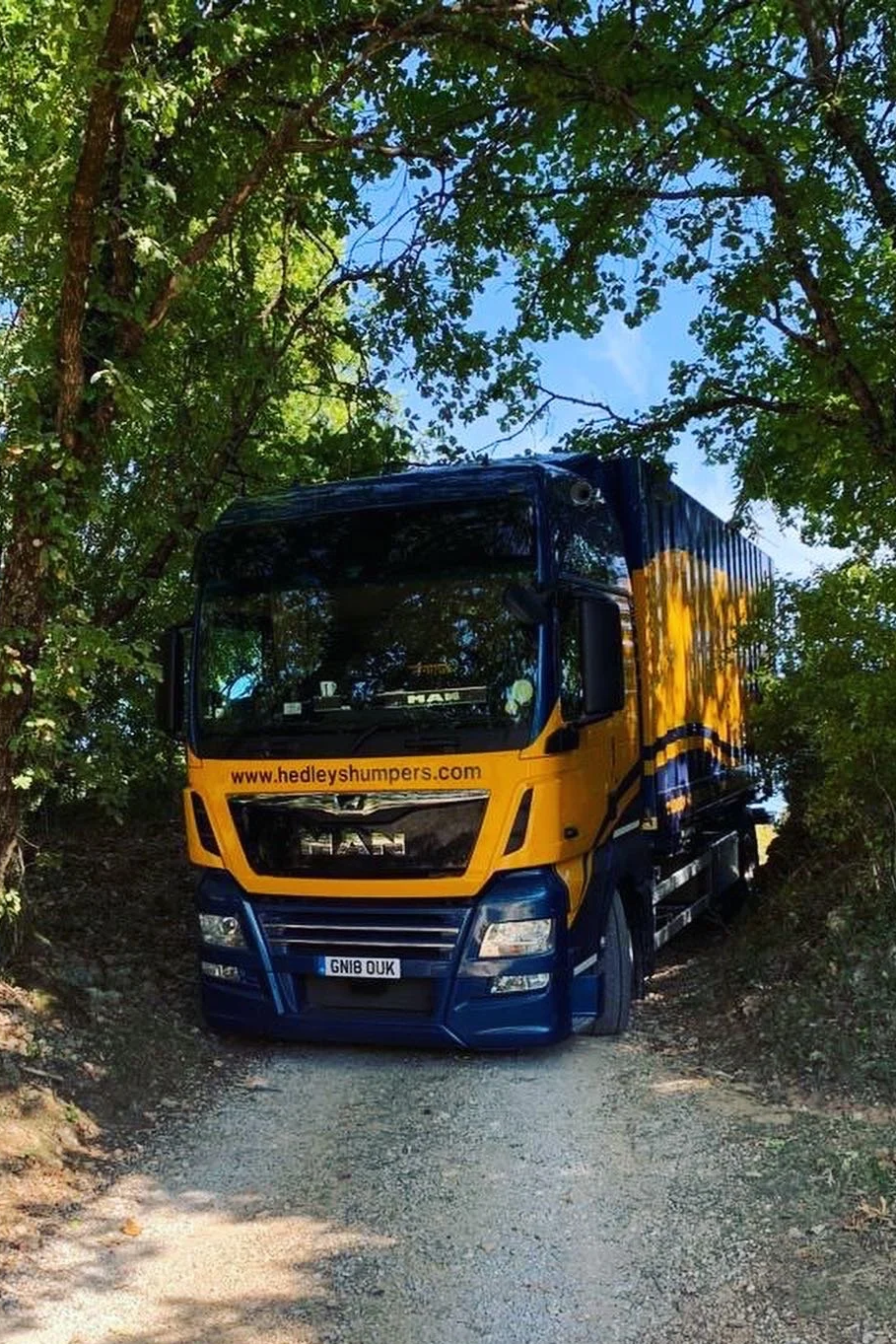 A large MAN truck painted in yellow and dark blue with the website www.hedleyshumpers.com on the front, parked on a dirt path surrounded by green trees and blue sky visible through the foliage.