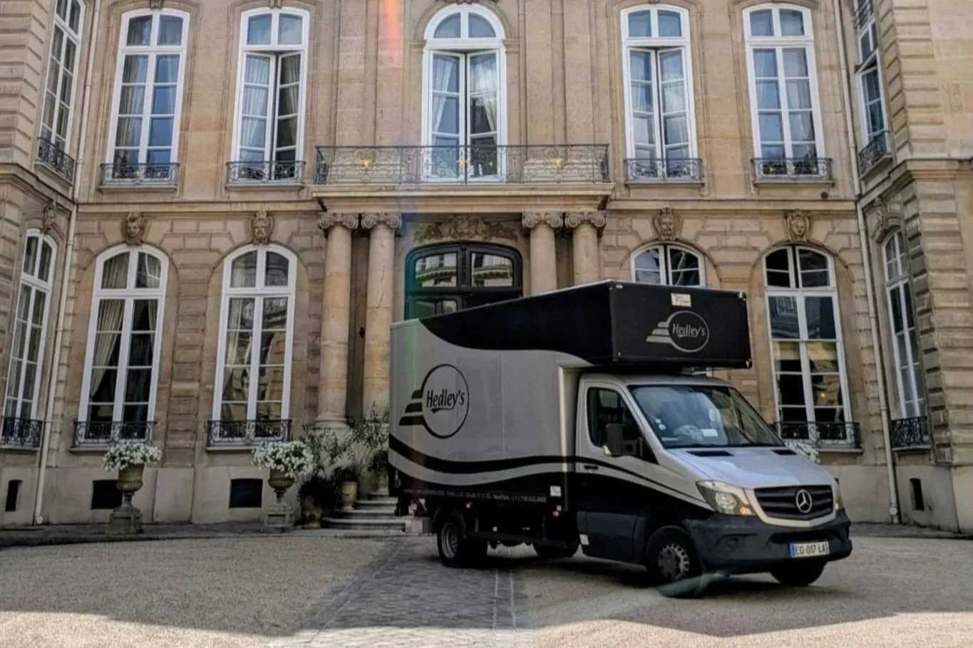 A delivery truck with Hedley's logo parked in front of an elegant, historic European-style building with large windows and ornate details.