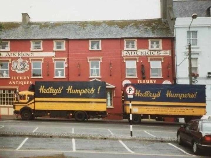 A street scene showing a two-story red building with the sign 'Fleury Antiques' and two trucks parked in front, both advertising 'Hedley's Humper!' in yellow text on a blue background.