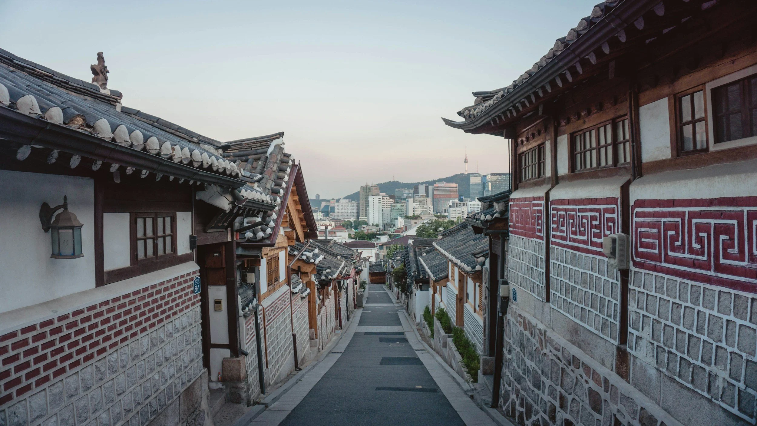 An alleyway between traditional Korean hanok houses with tiled roofs, leading to a modern cityscape with high-rise buildings in the background.