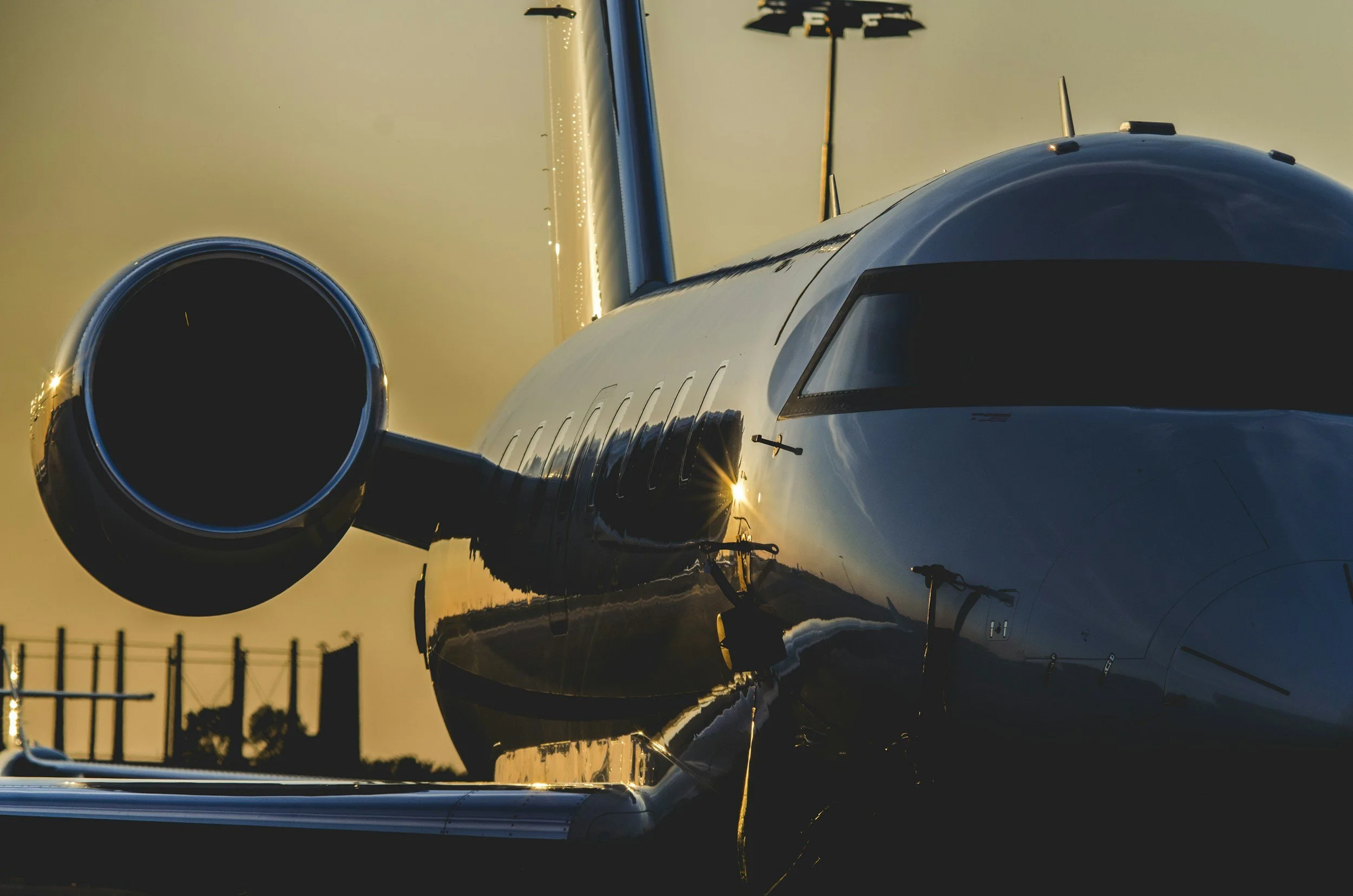 Close-up of a private jet aircraft at sunset, showing the front portion of the fuselage, the cockpit windows, and one engine on the left side, with the sun reflecting off the plane's shiny surface.