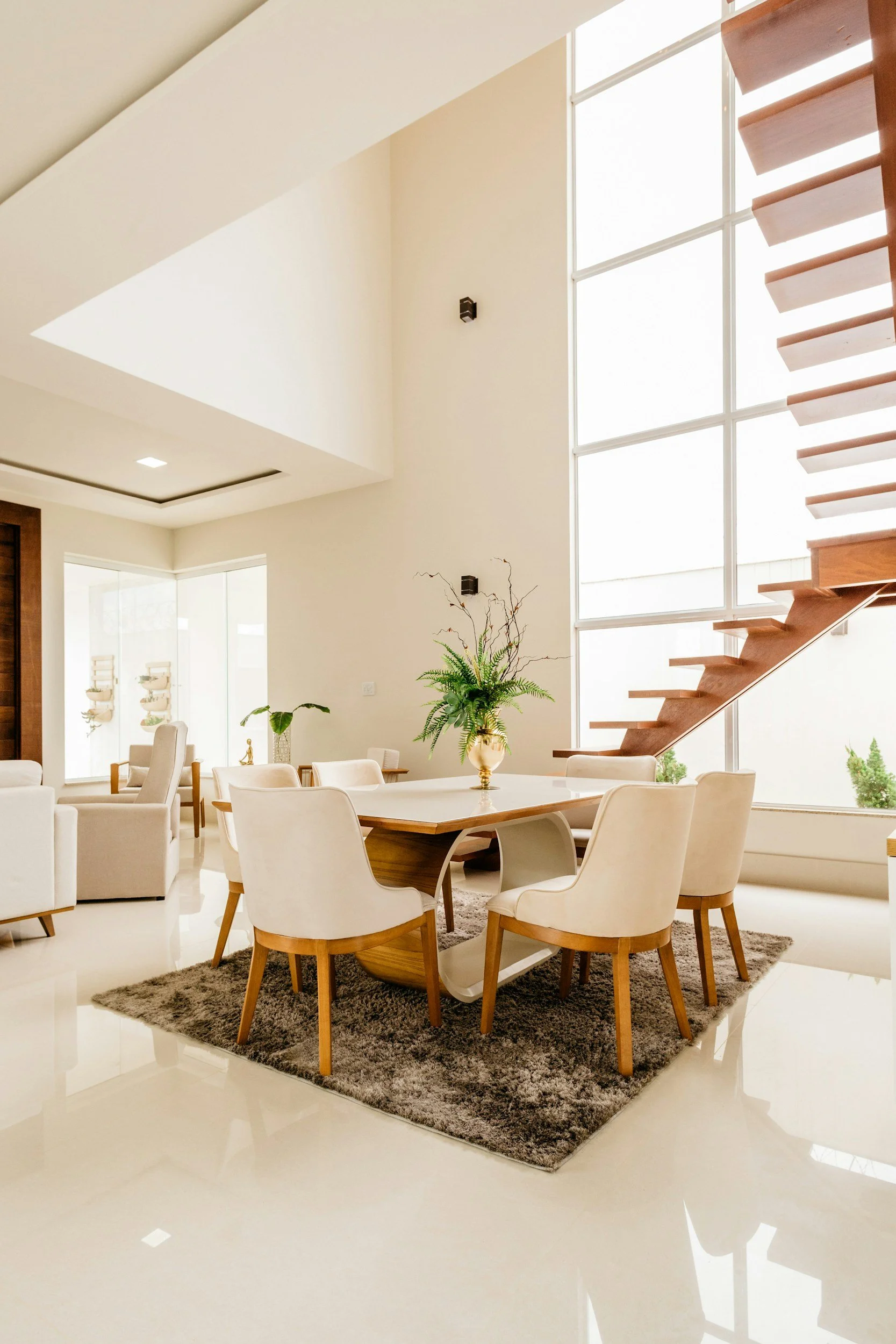 Modern dining area with a large glass window, white chairs around a wooden table on a gray rug, and green plants nearby. Wooden staircase ascending along the window.