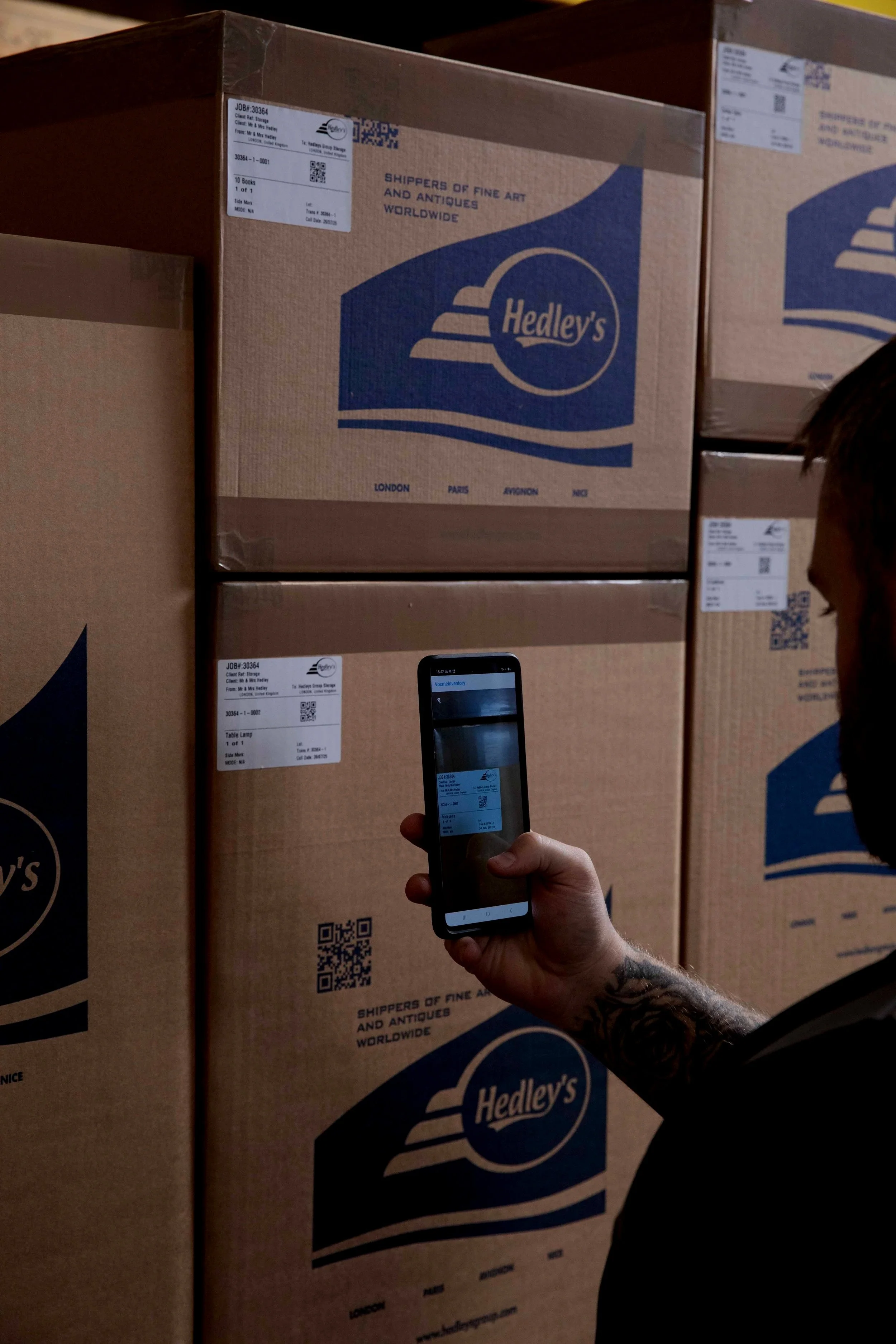 Person in a black shirt holding a smartphone, taking a photo of large cardboard boxes labeled Hedley's, in a warehouse or storage area.