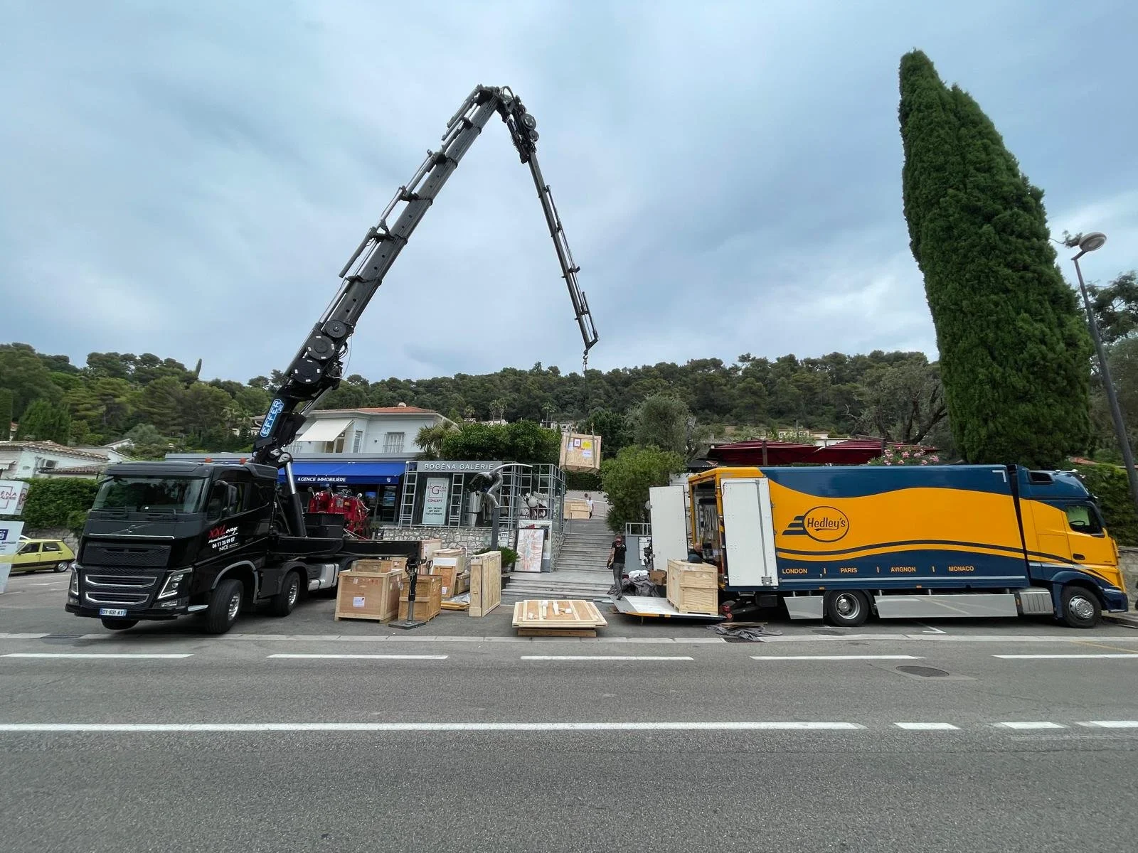 A delivery truck with a mounted crane unloading wooden crates in front of a gallery store on a city street.