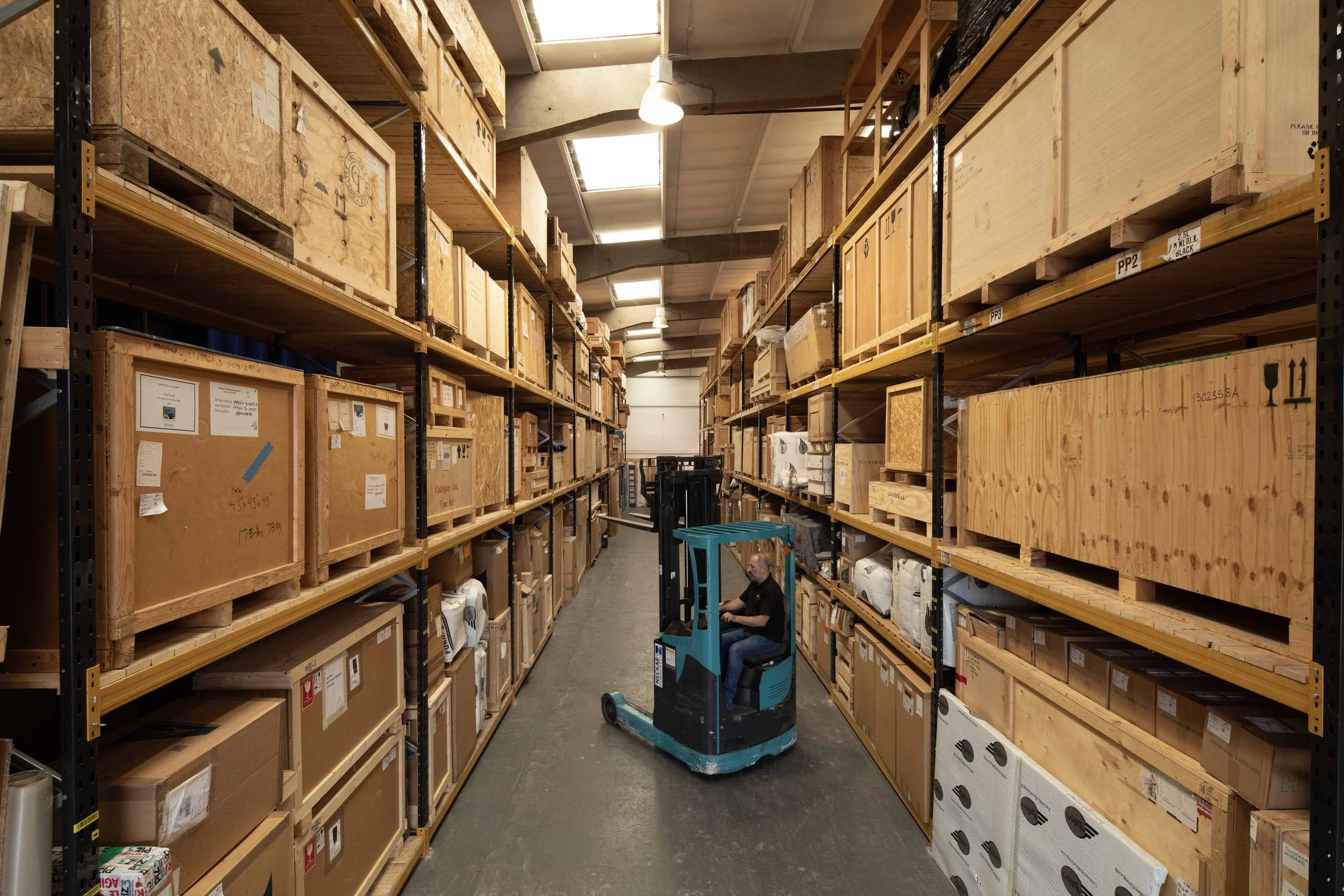 A warehouse worker operating a forklift among tall shelves filled with wooden crates and boxes.