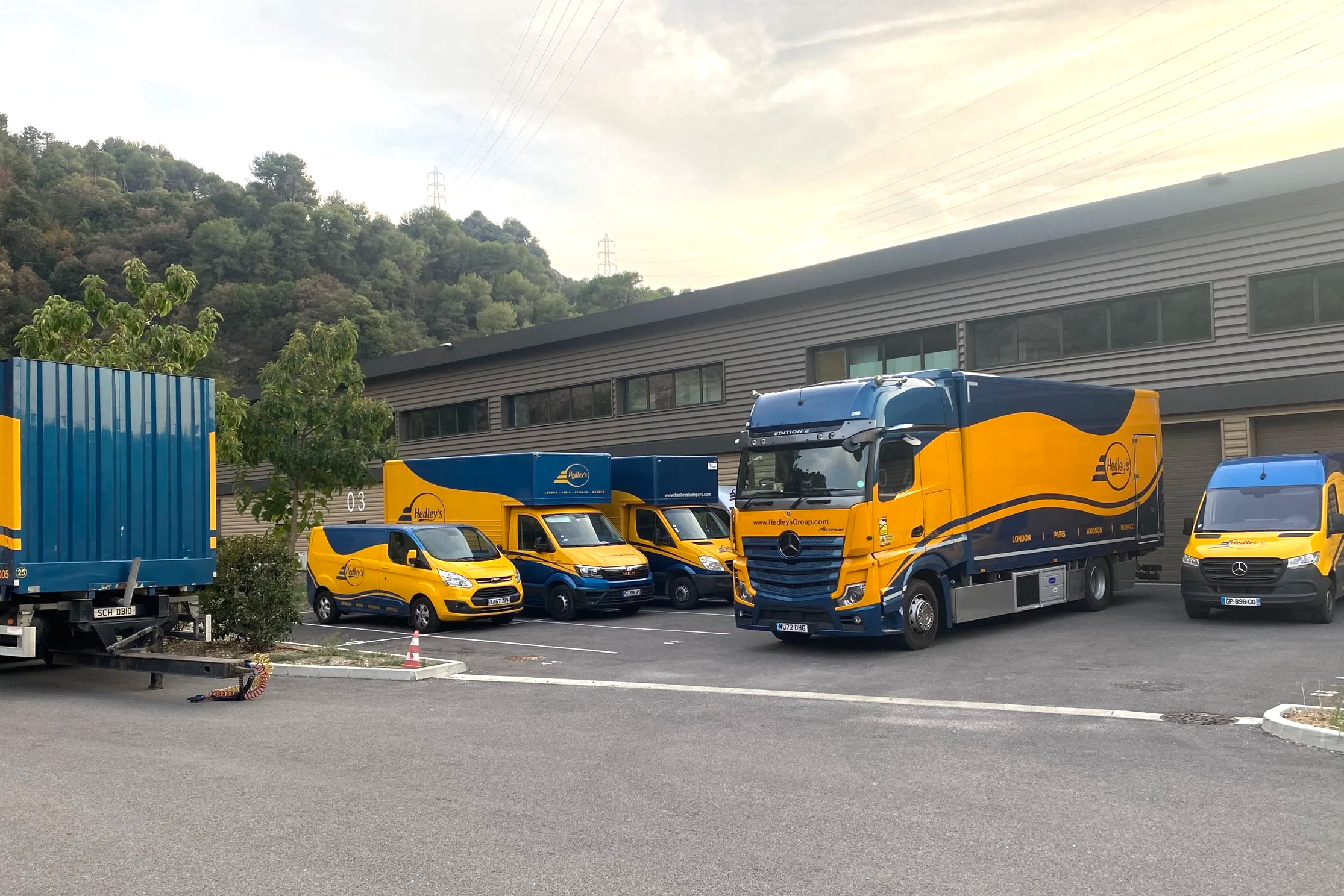 A parking lot with several yellow and blue delivery trucks and vans with Hedley's branding, positioned near a gray industrial building and a green hillside.