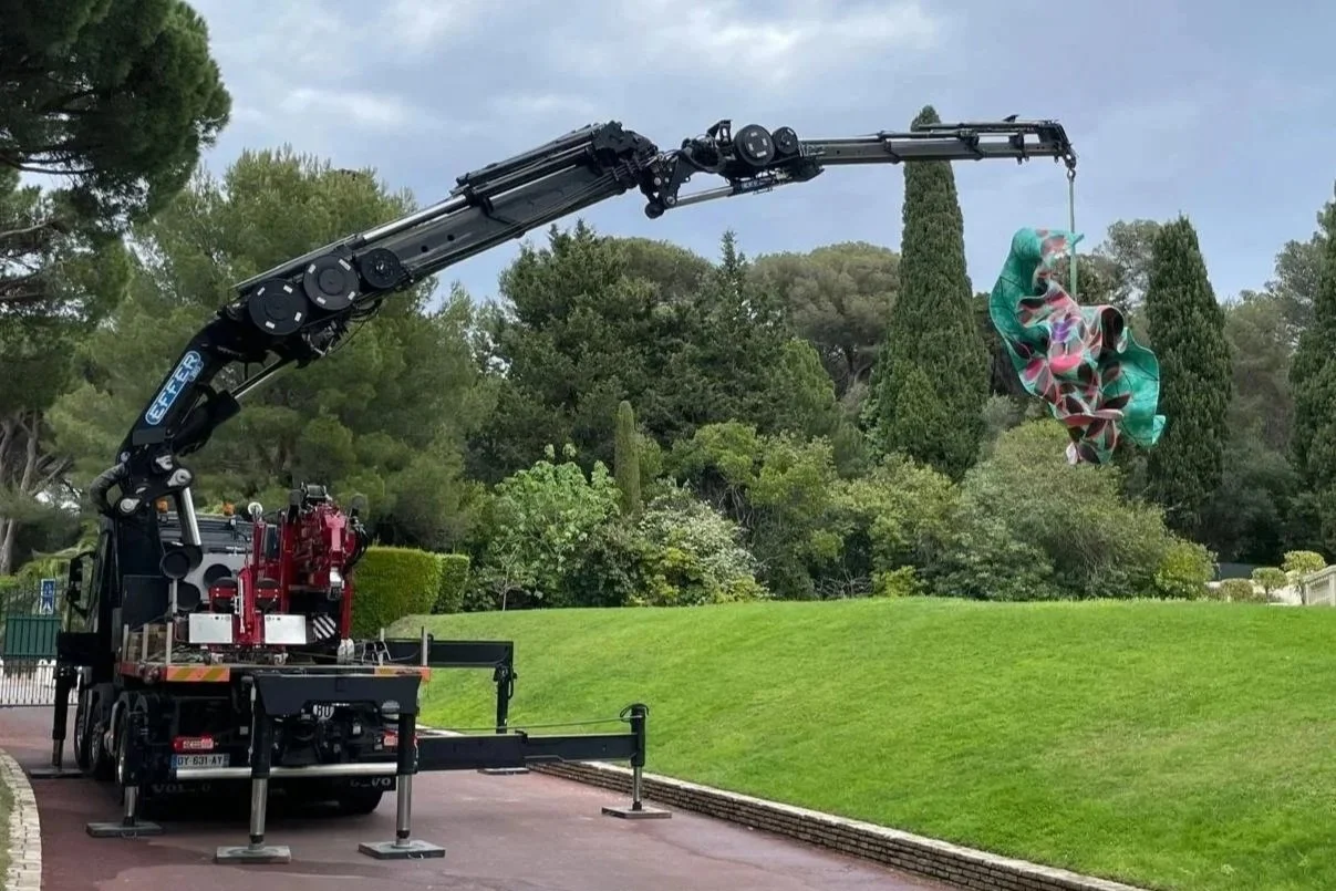 Mobile crane unloading large wooden crate from a Hendley's branded delivery truck on a street, with a hillside and cloudy sky in the background.