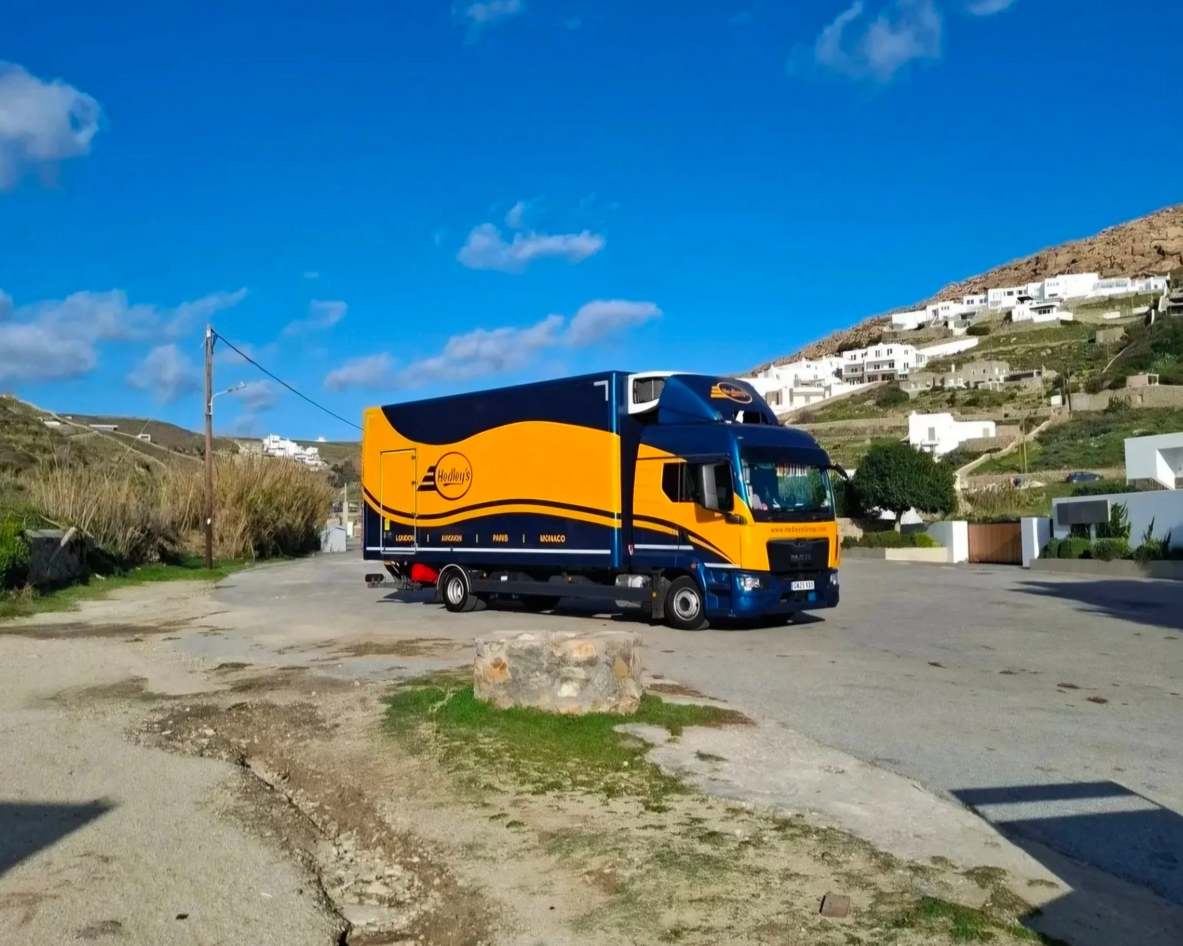 Aerial view of a picturesque coastal town with colorful buildings, a marina with boats, an island with trees, and mountainous landscape in the background.