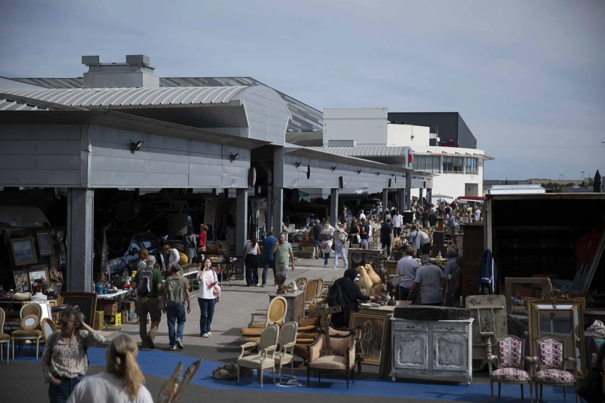 A busy outdoor flea market with vendors selling furniture and other items, and many people browsing and walking through the area.