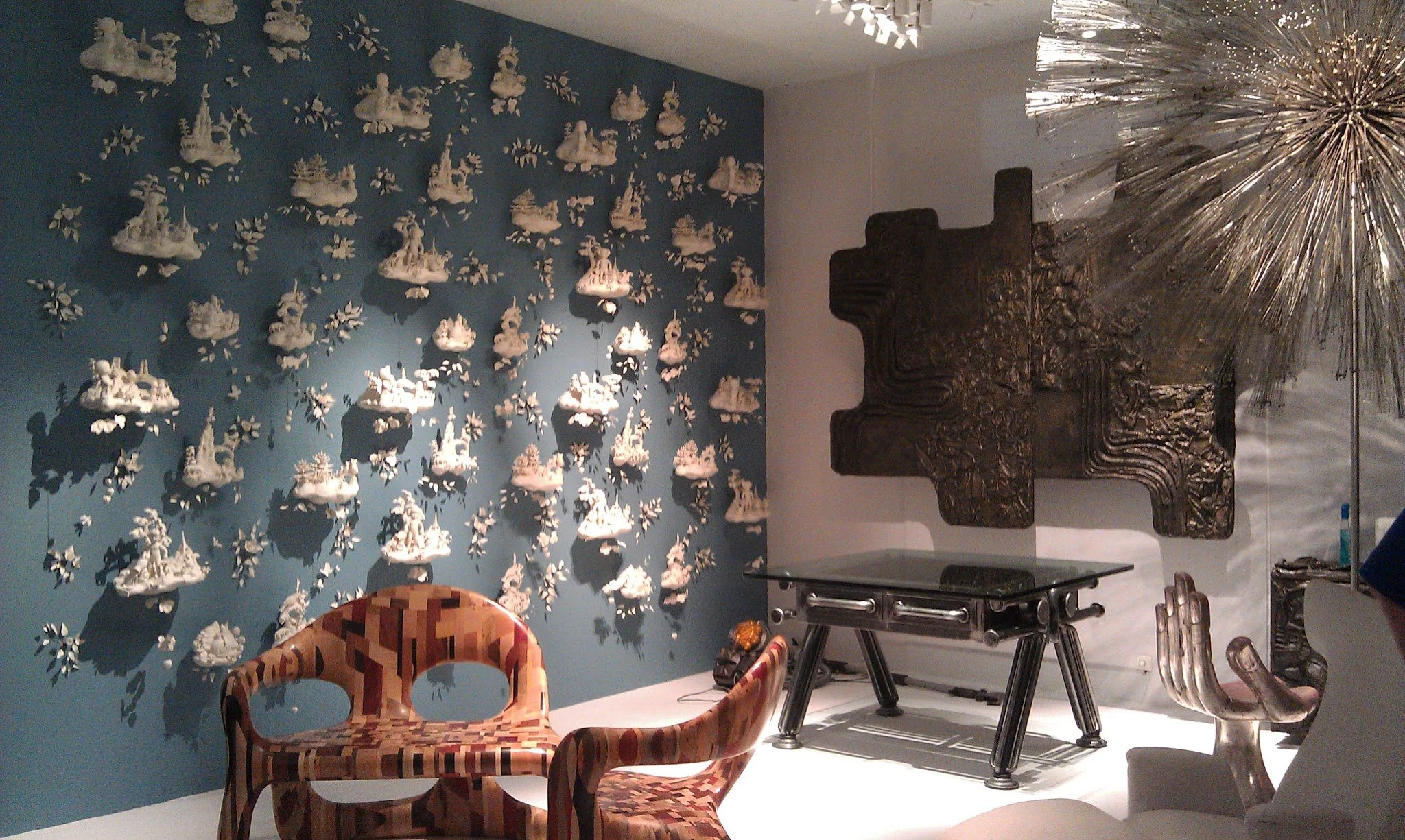 An art gallery wall with white cloud sculptures casting shadows, a dark textured wall sculpture, and modern furniture including a colorful patterned armchair and a glass table.