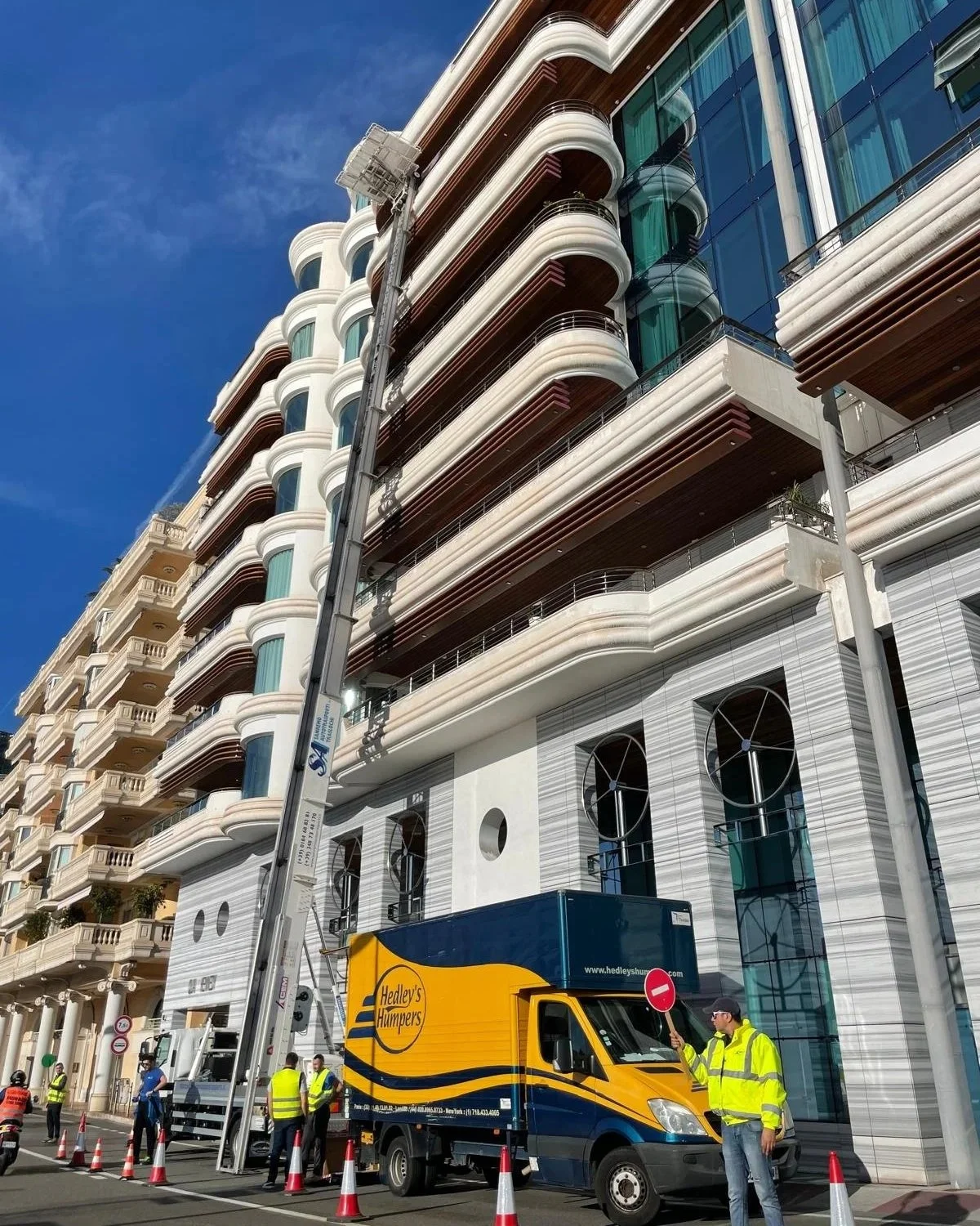 Construction workers and a Hedley's Humbugs truck in front of a modern multi-story building with curved balconies and large glass windows.