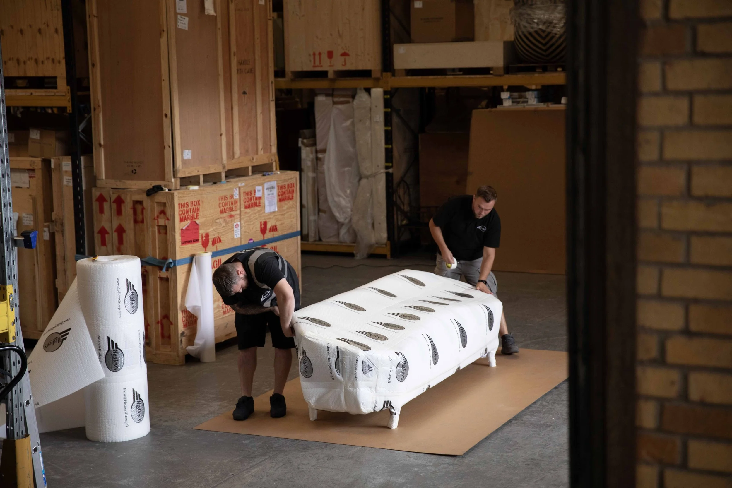 Two workers are wrapping a sofa with protective plastic in a warehouse storage area, surrounded by large cardboard boxes and shelves.