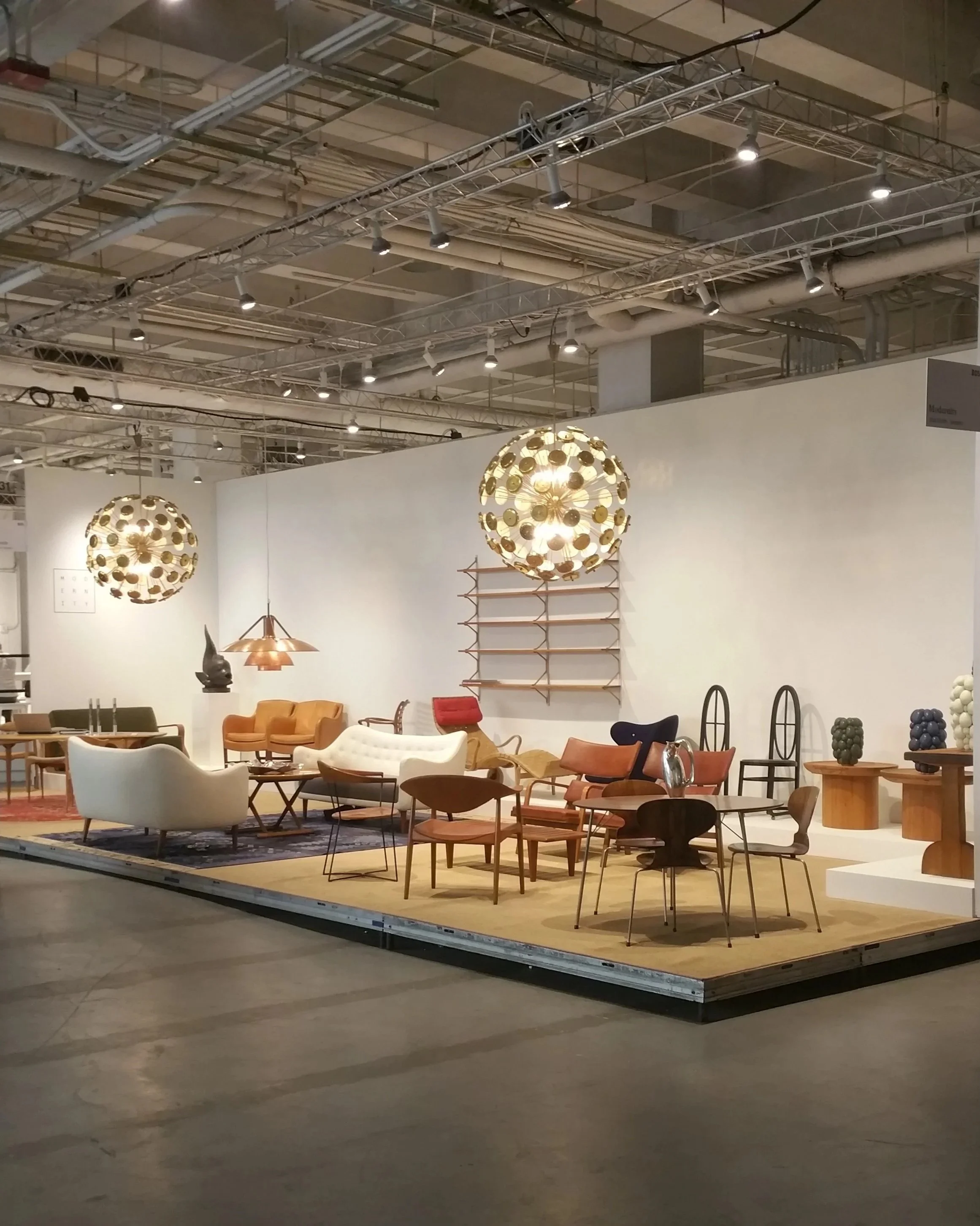 An indoor furniture display featuring various chairs, sofas, and tables on a beige rug, with decorative lighting fixtures hanging from the ceiling, and a white wall background with shelving and decorative objects.