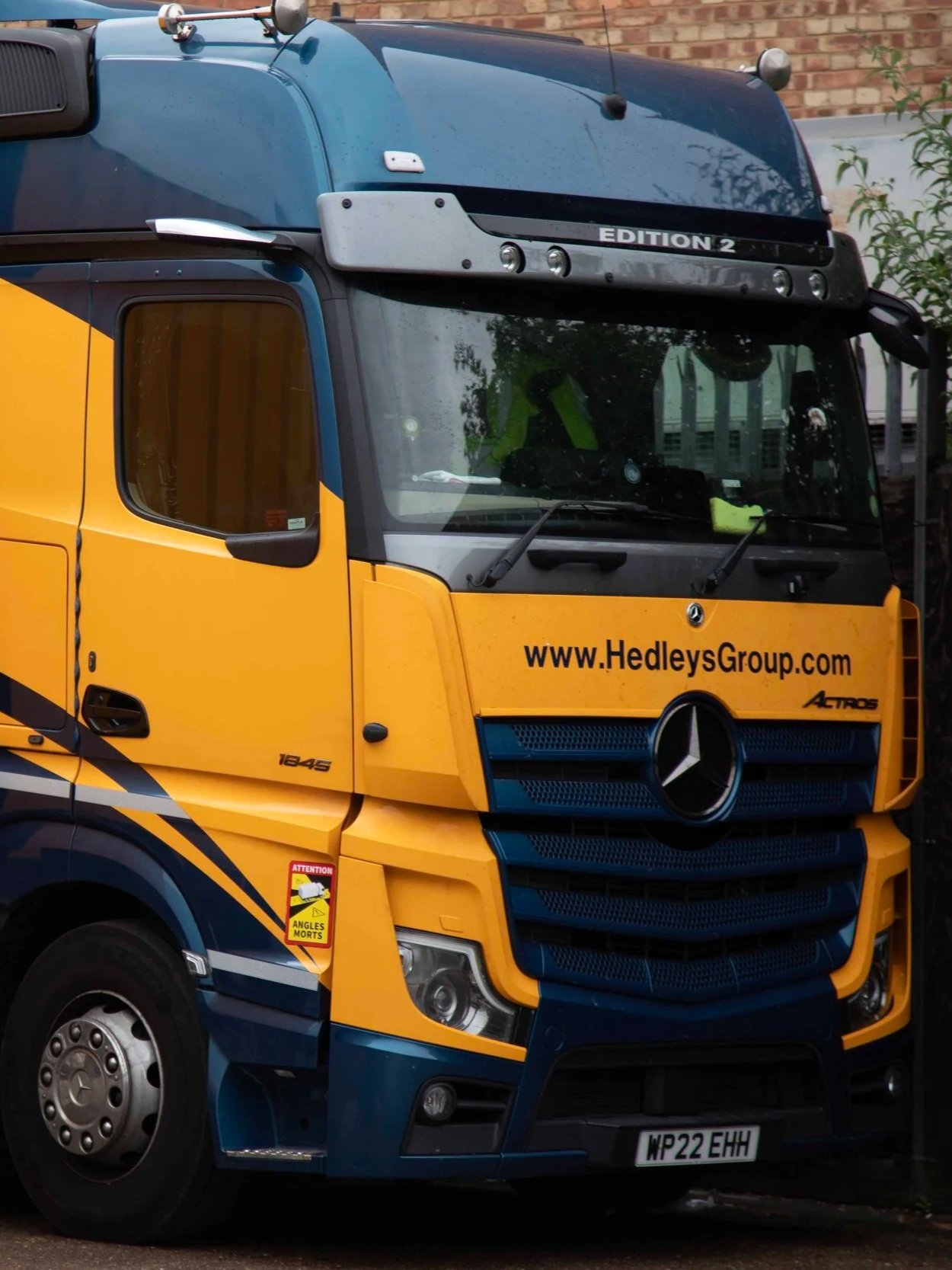 Yellow and blue Mercedes-Benz truck with website www.HedleysGroup.com on the front, license plate WP22 EHH, and a warning sign that says 'Attention Angles Morts' on the side, parked in front of a brick wall.