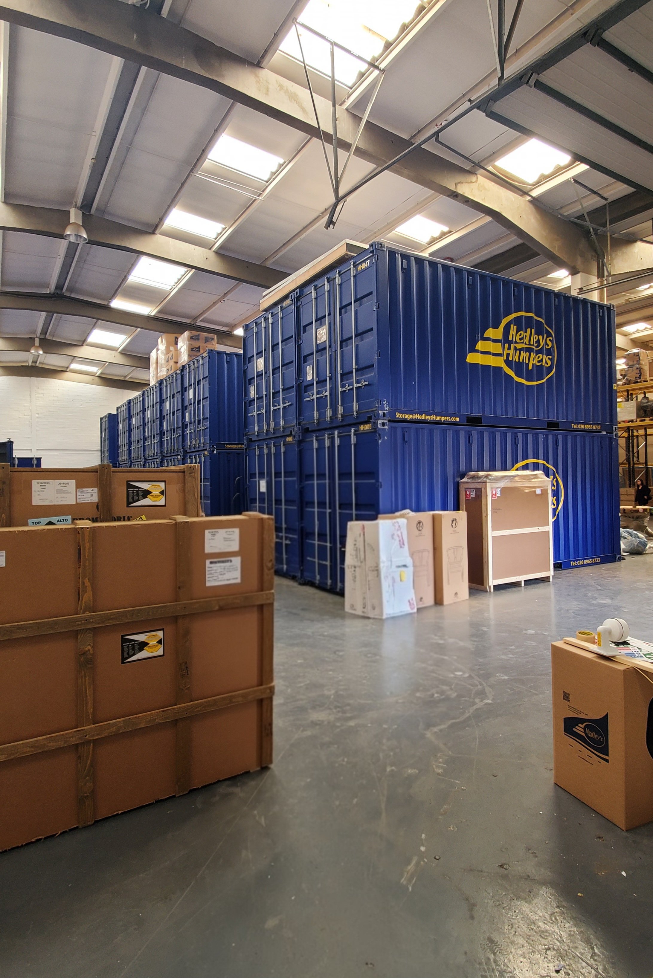 Warehouse interior with stacked blue shipping containers labeled Hedley's Humpeys, and brown cardboard boxes on the floor.