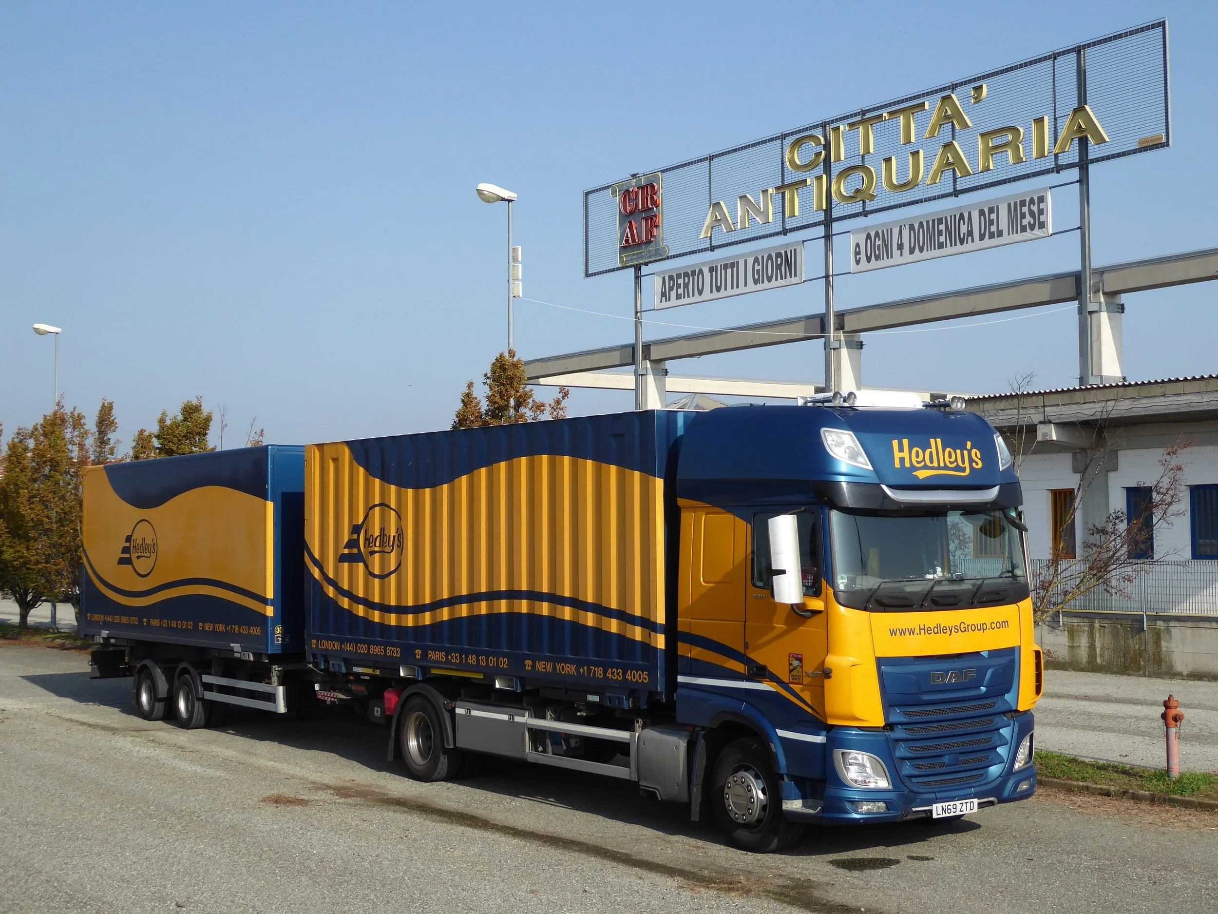 A Hesley's delivery truck parked on the street near a sign that reads 'Città Antiquaria' and 'Aperto Tutti i Giorni', with trees and a building in the background.