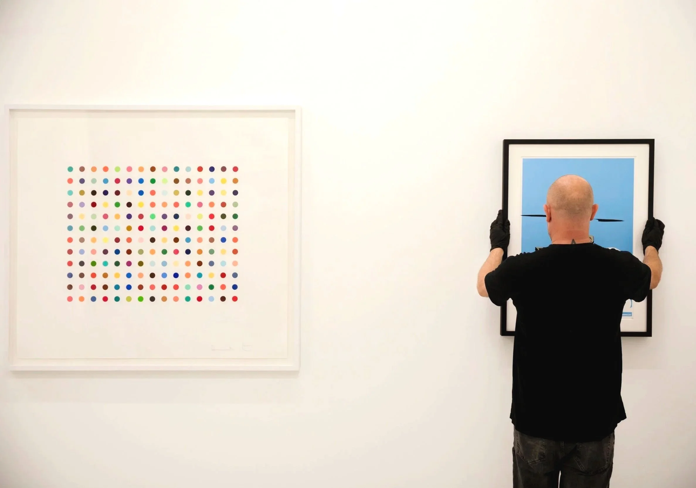 An art gallery wall with white cloud sculptures casting shadows, a dark textured wall sculpture, and modern furniture including a colorful patterned armchair and a glass table.