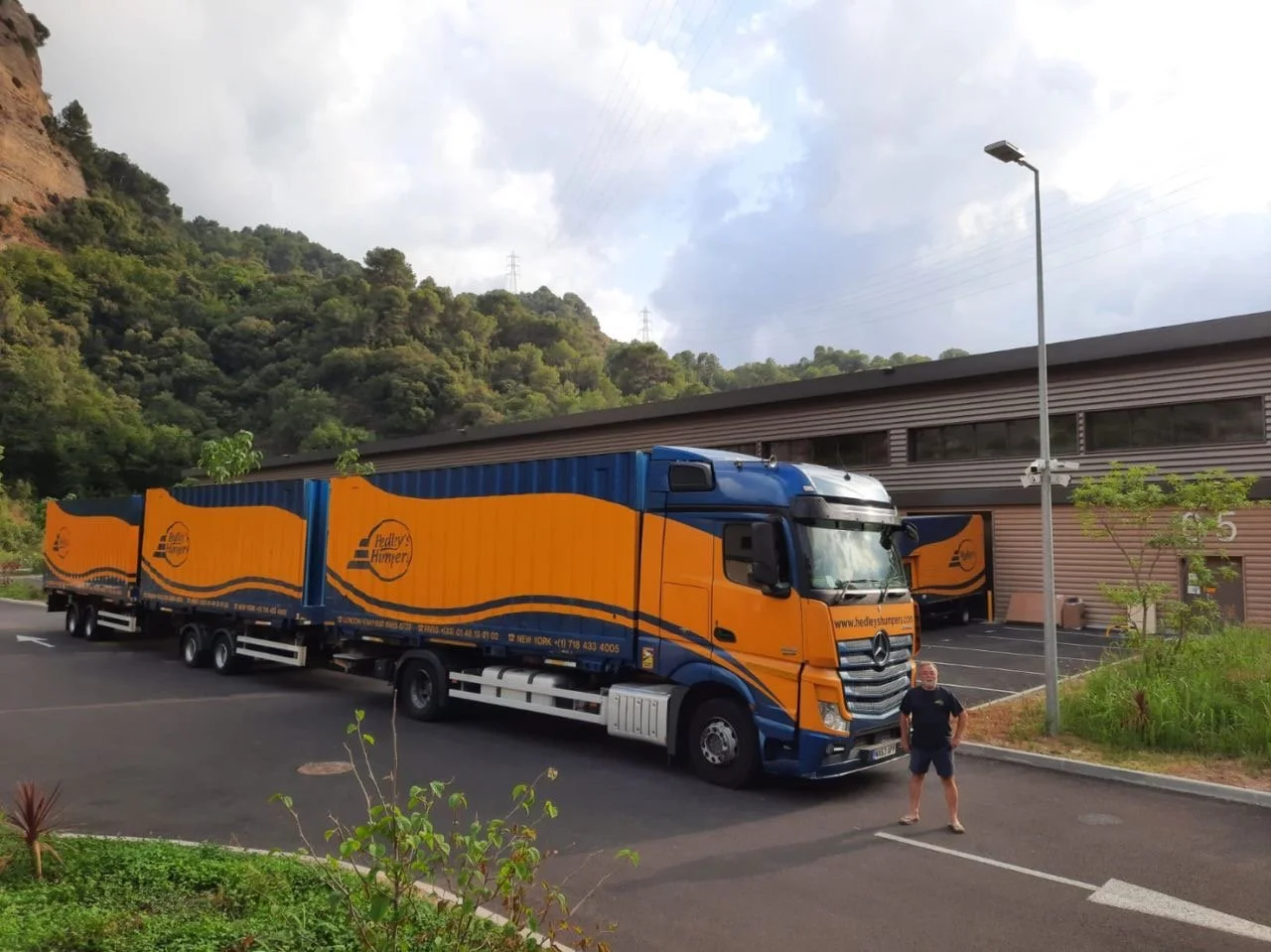 A man stands next to a large orange and blue truck parked in front of a building with a hillside covered in trees behind it. There is a streetlamp and some small plants in the foreground.