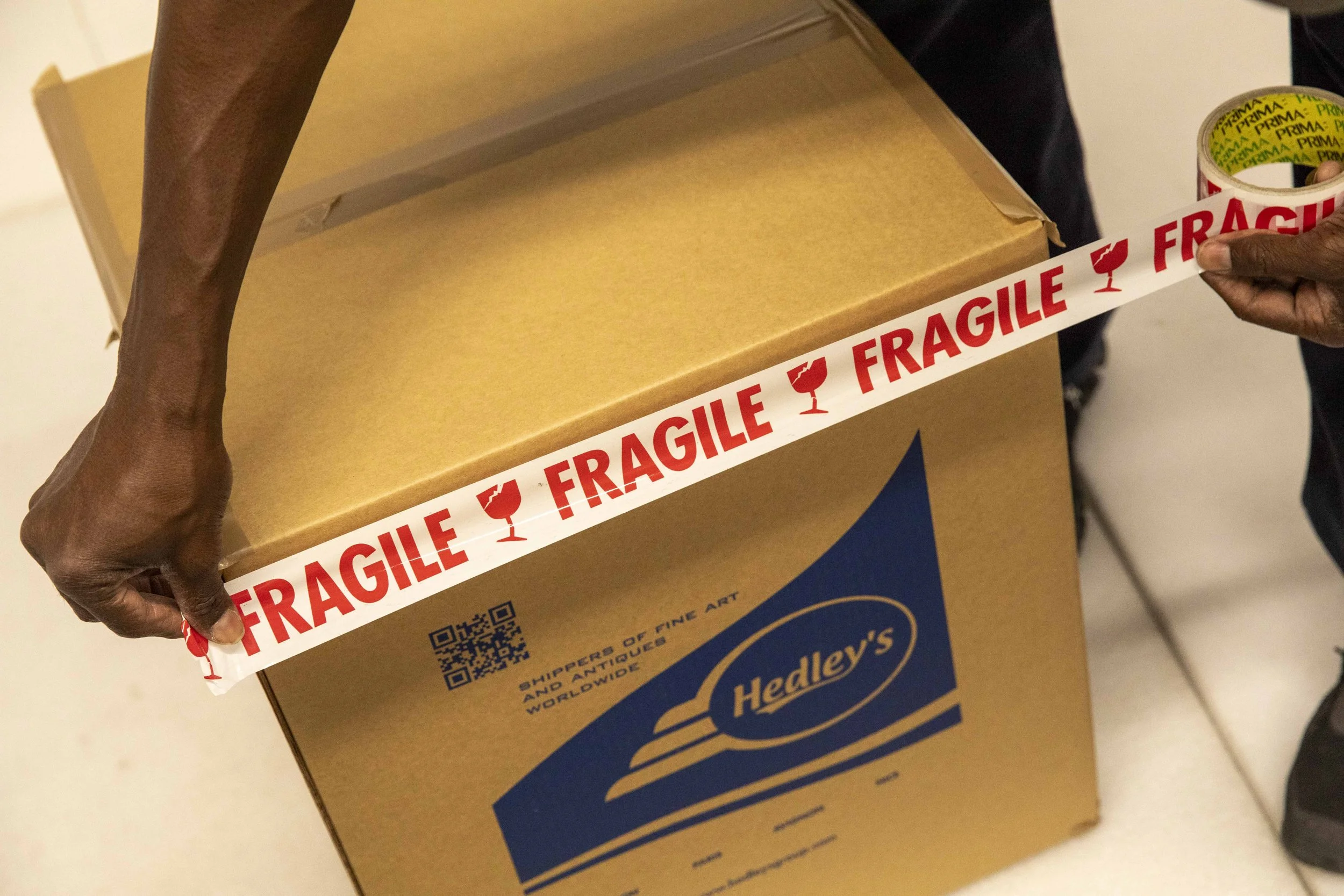 A person is sealing a box with red 'Fragile' tape labeled with a wine glass icon. The box is cardboard with a blue logo for Hedley's and a QR code. The person has dark skin and is wearing a black shirt.