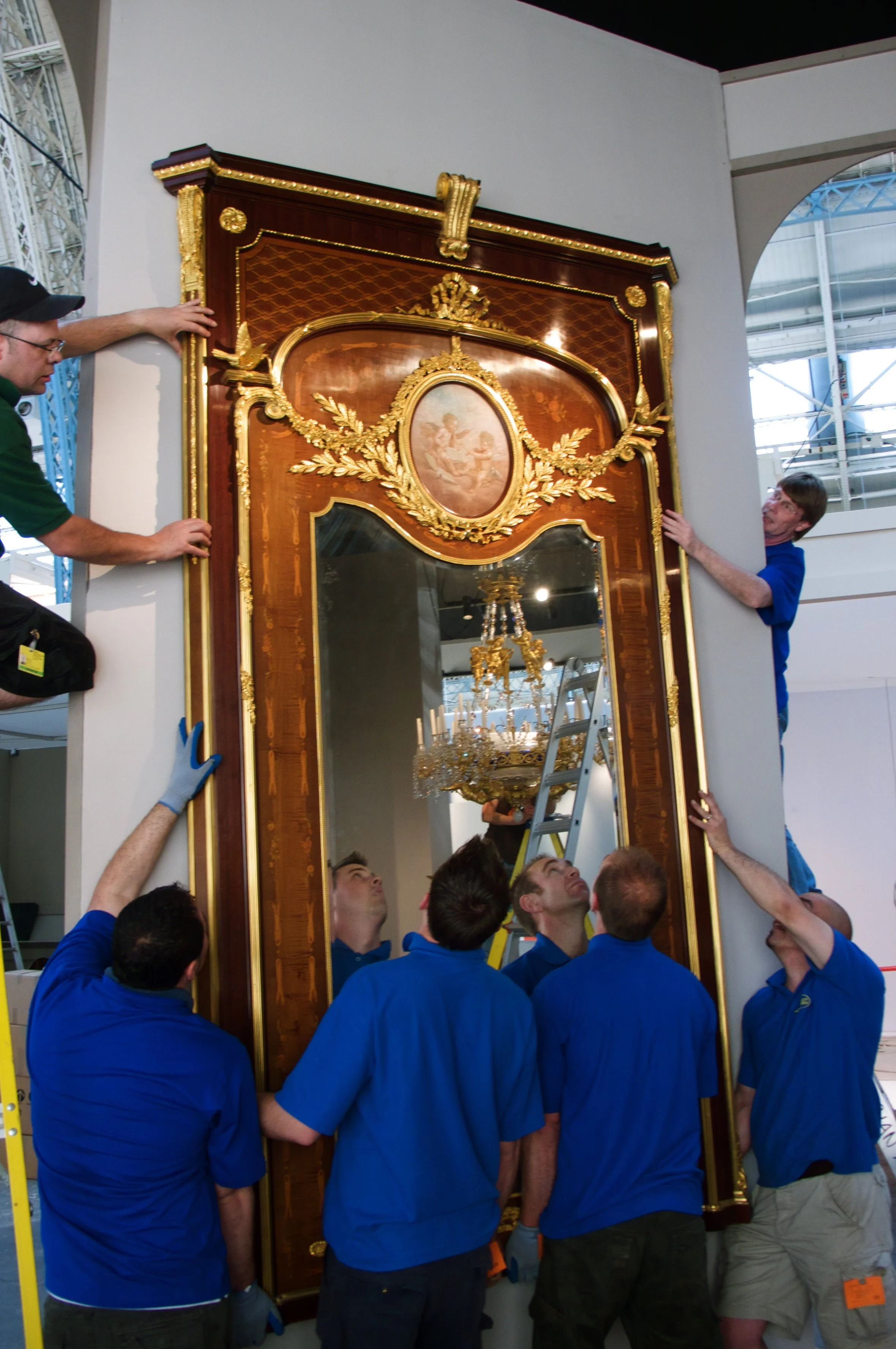 Group of people carefully installing a large, ornate gilded mirror with a painted oval portrait at the top and decorative gold accents, in an industrial-style setting.