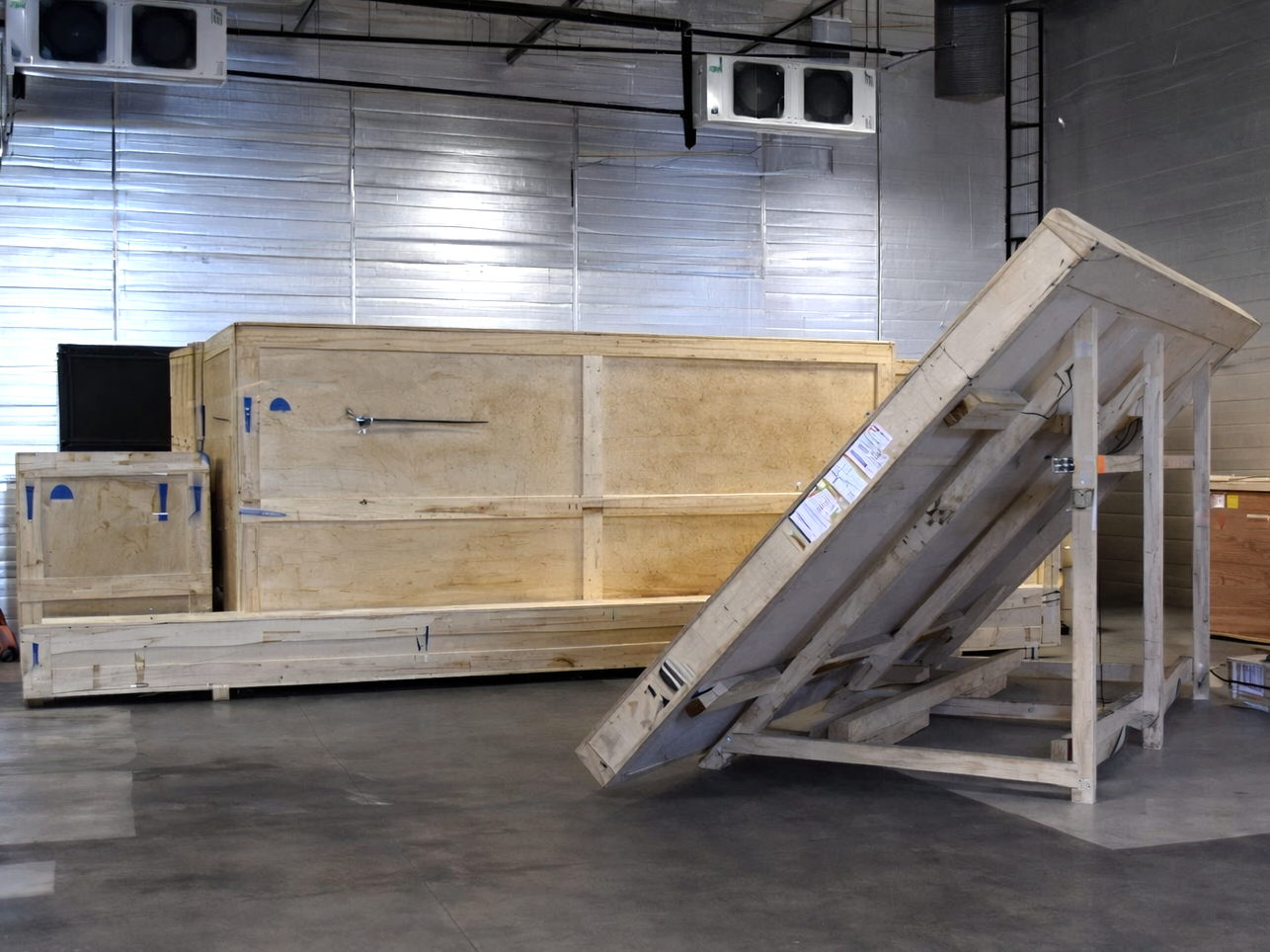 Wooden crates and pallets inside a warehouse with metallic walls and ceiling-mounted air conditioning units.