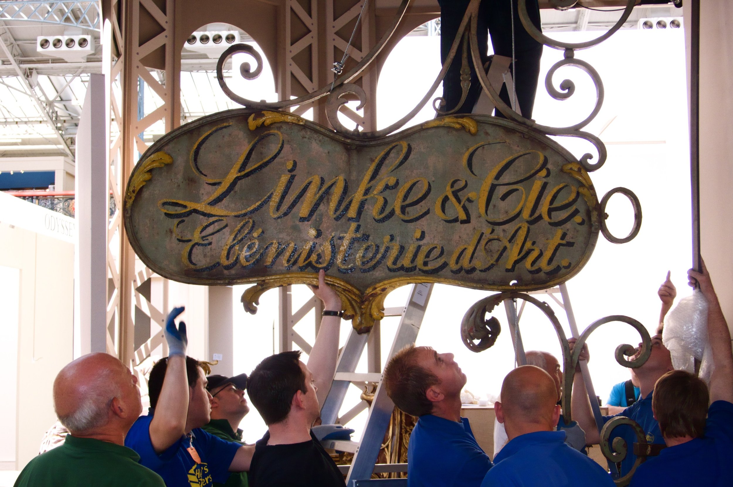 Group of people installing or maintaining a vintage sign that reads 'Limke & Cie, Ébénisterie d'Art' in a building with a high ceiling and industrial lighting.
