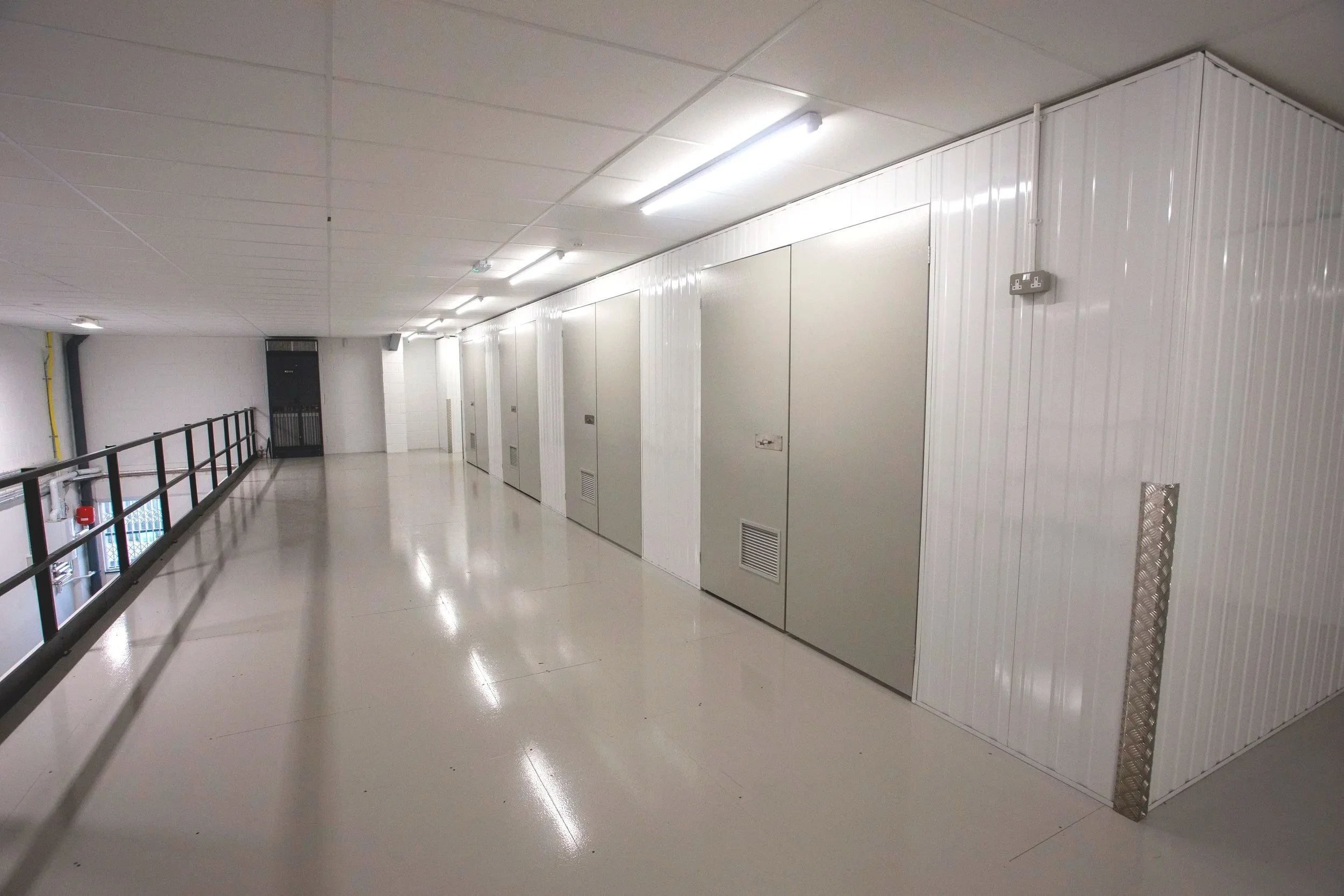 Empty industrial hallway with white walls, ceiling, and floor, featuring multiple closed compartments or electrical cabinets along the wall, fluorescent lighting, and a safety railing on the left side.