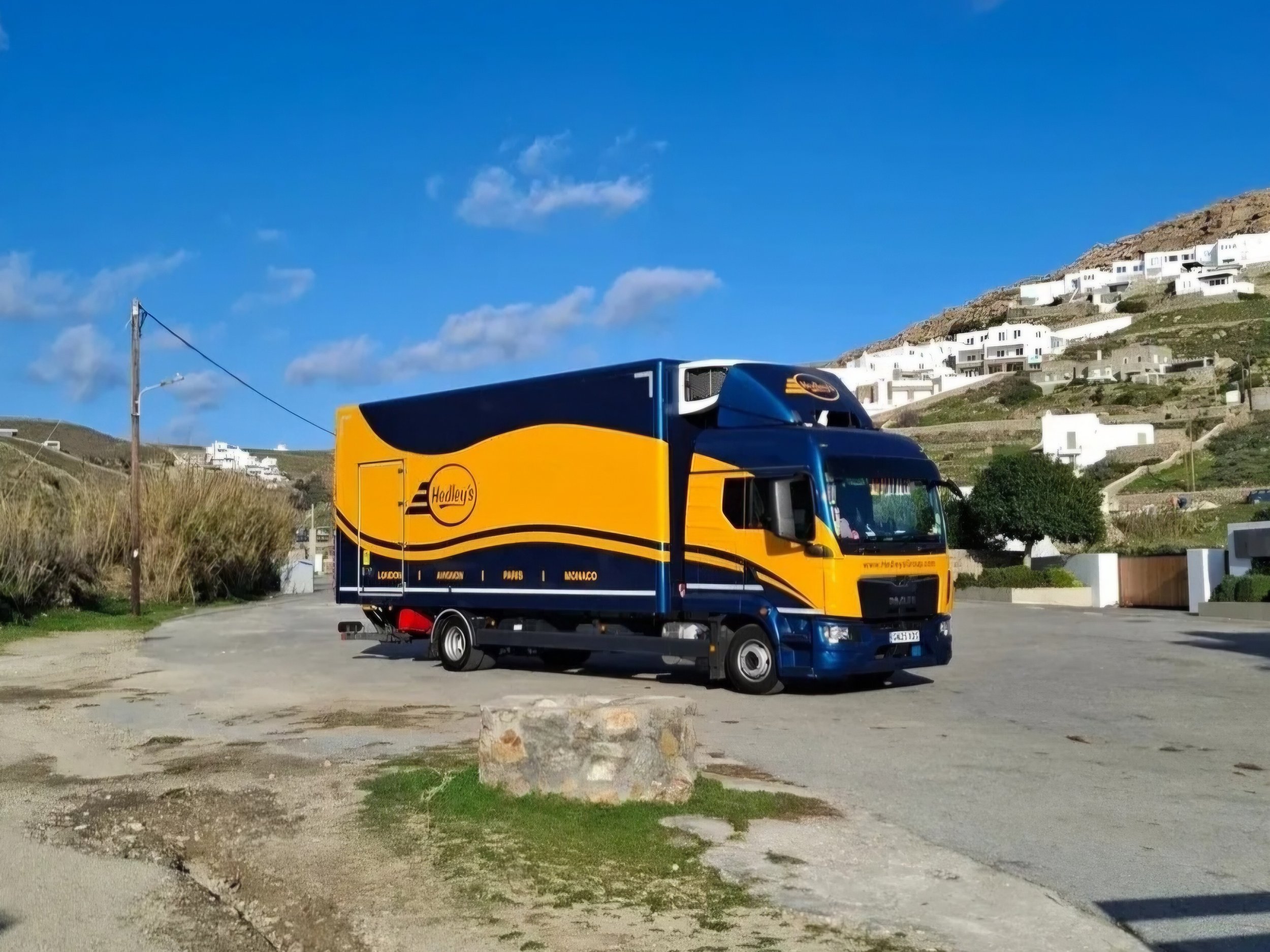 Aerial view of a picturesque coastal town with colorful buildings, a marina with boats, an island with trees, and mountainous landscape in the background.
