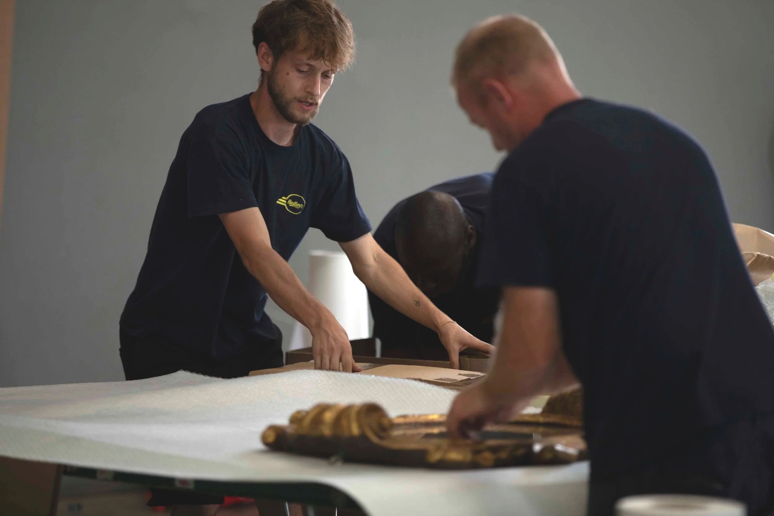Three men preparing a large sheet of material on a table, with some baked goods on a tray in the foreground, in a room with gray walls.