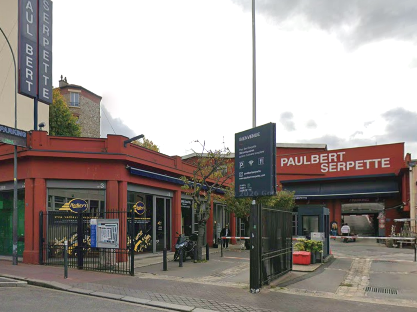 A street view showing a red building with antique clocks inside a shop called Hedley's, and a man wearing a gray hoodie walking past holding a cardboard piece. There are parked motorcycles on the sidewalk, a fenced area with trees, and adjacent buildings with signs reading 'Serpette' and 'Julbert Serpette'.