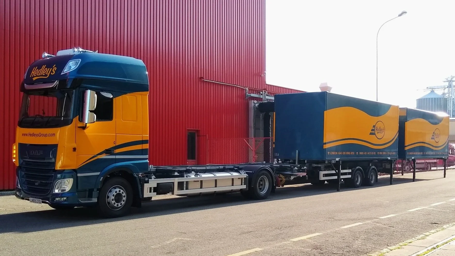A yellow and dark blue truck with Hedley's branding parked on the street next to a red building, with large blue trailers attached.