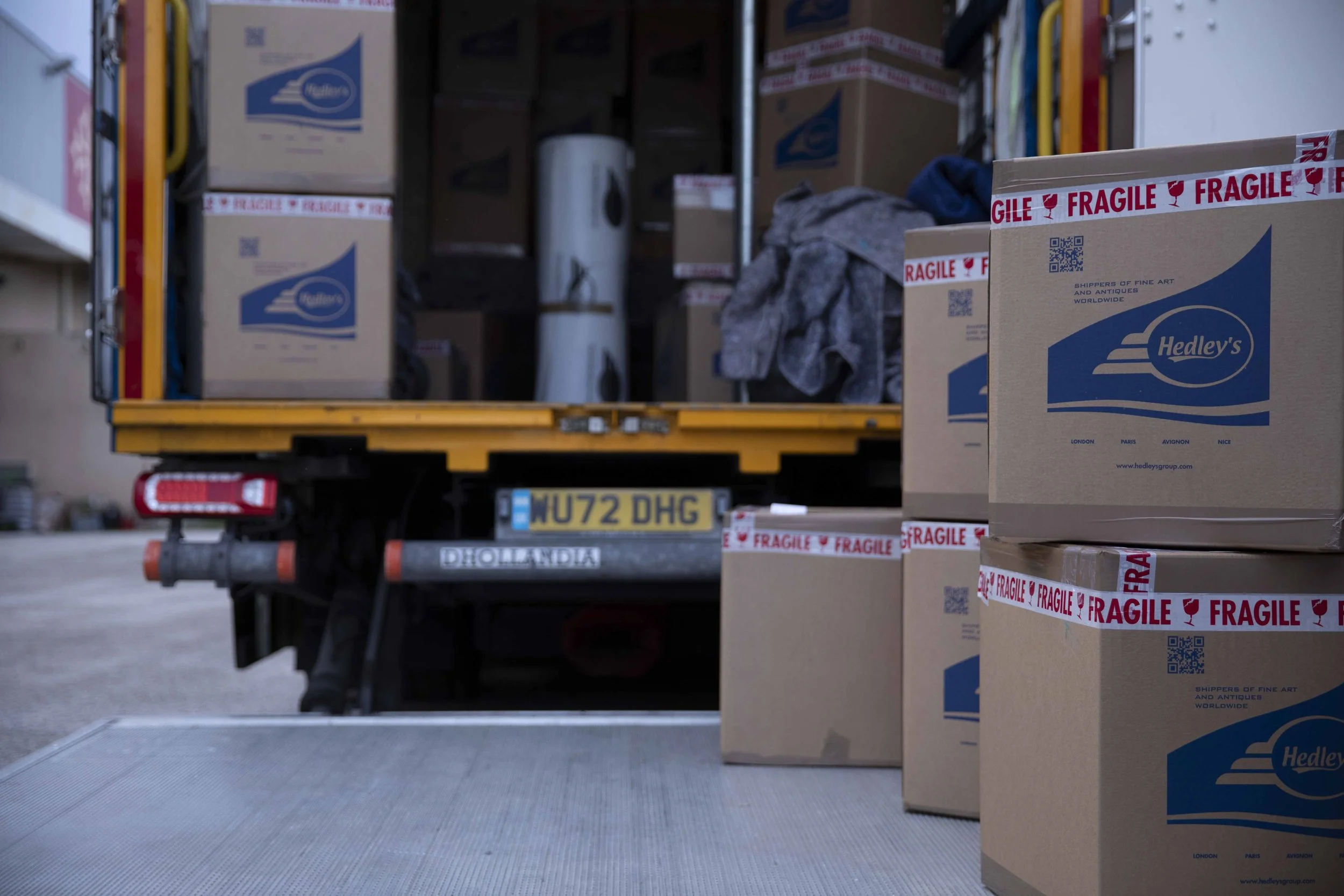 A yellow delivery truck with an open back filled with boxes and packages. Several cardboard boxes are in the foreground, some labeled 'Hedley's' with a blue logo and marked 'fragile' with red tape. The truck's license plate reads 'MU72 DHG' and the rear bumper has a 'Hollandia' sticker.