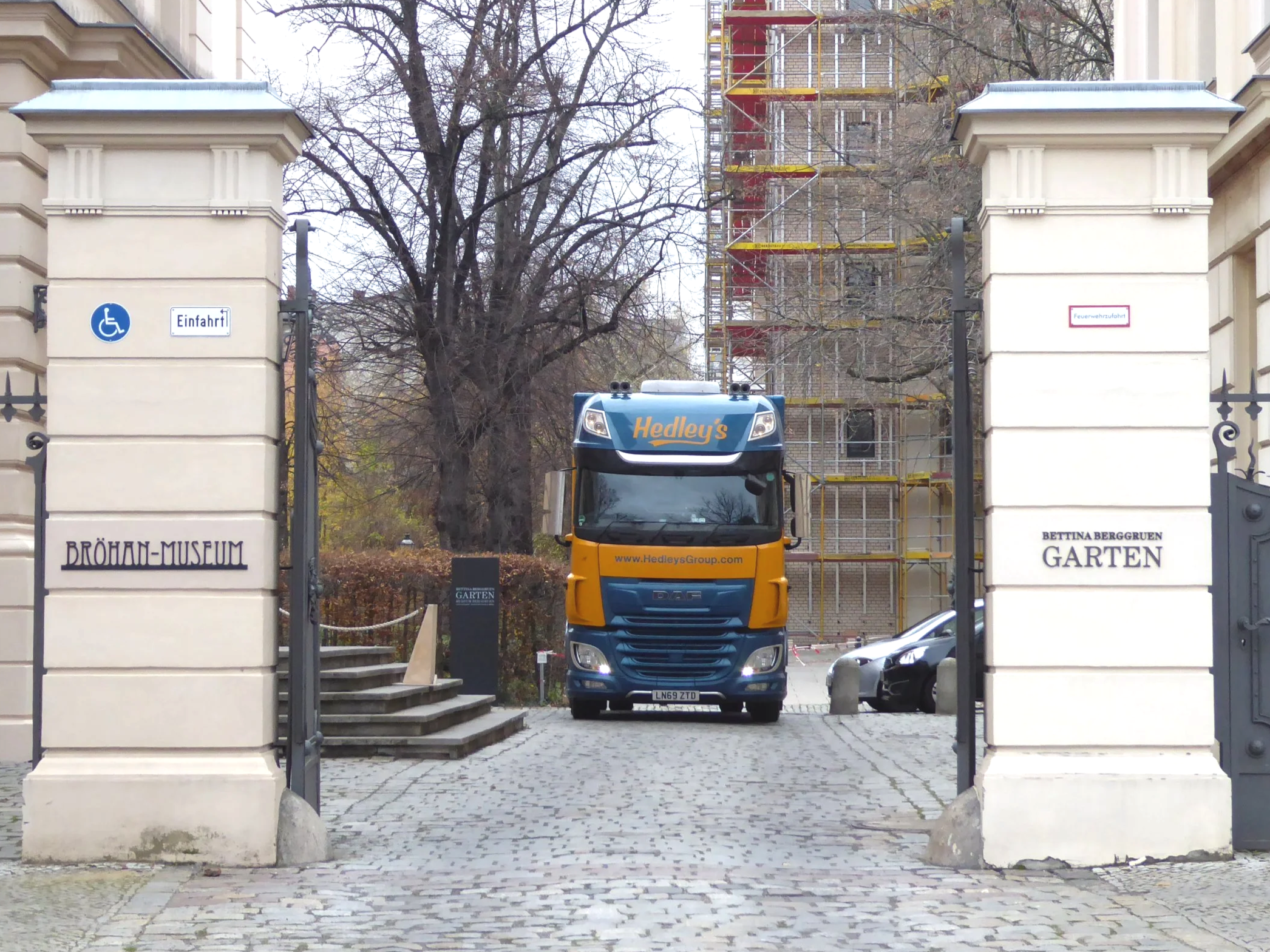 Entrance gate to the Bröhan Museum with a yellow and blue Hedley's Group truck in the background, stone pillars with signs, parked cars, leafless trees, and a building under construction with scaffolding.