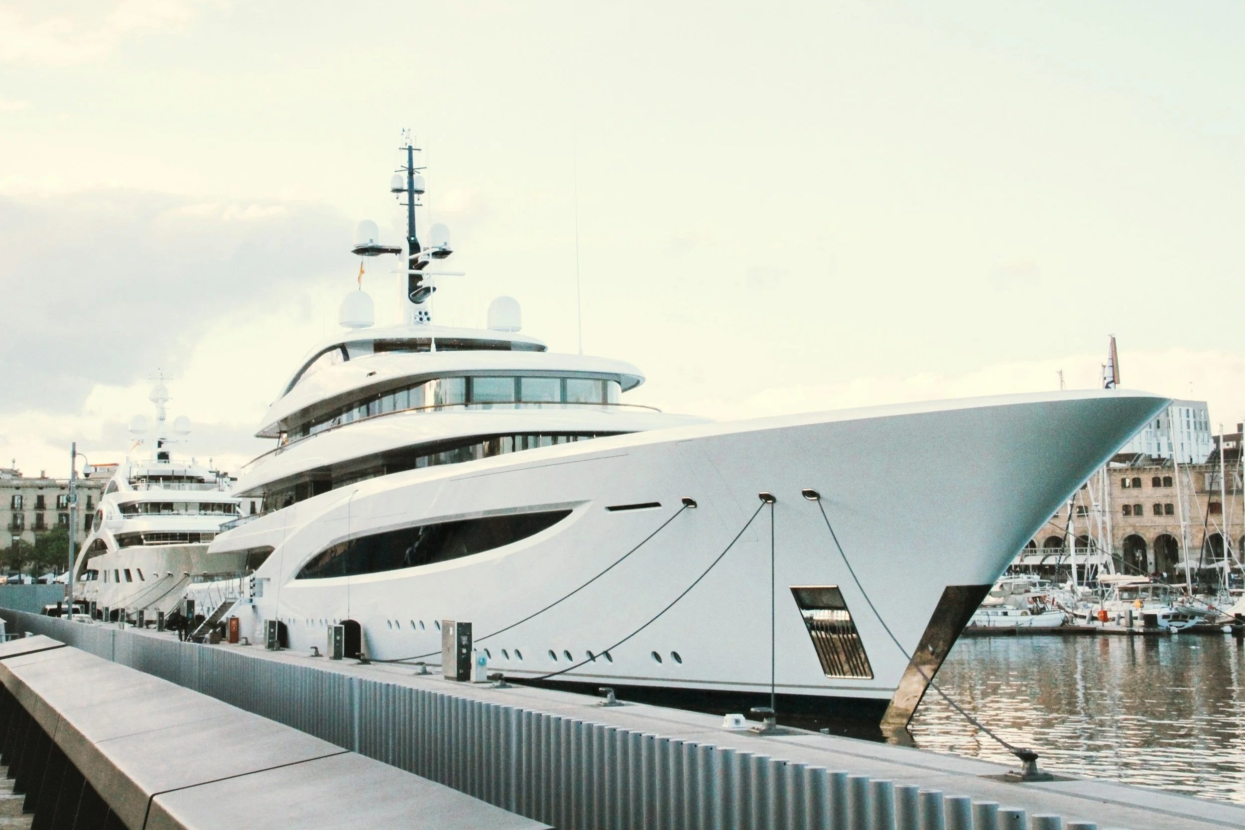 A large, modern yacht docked at a marina under a clear blue sky, with a flag flying on the stern.